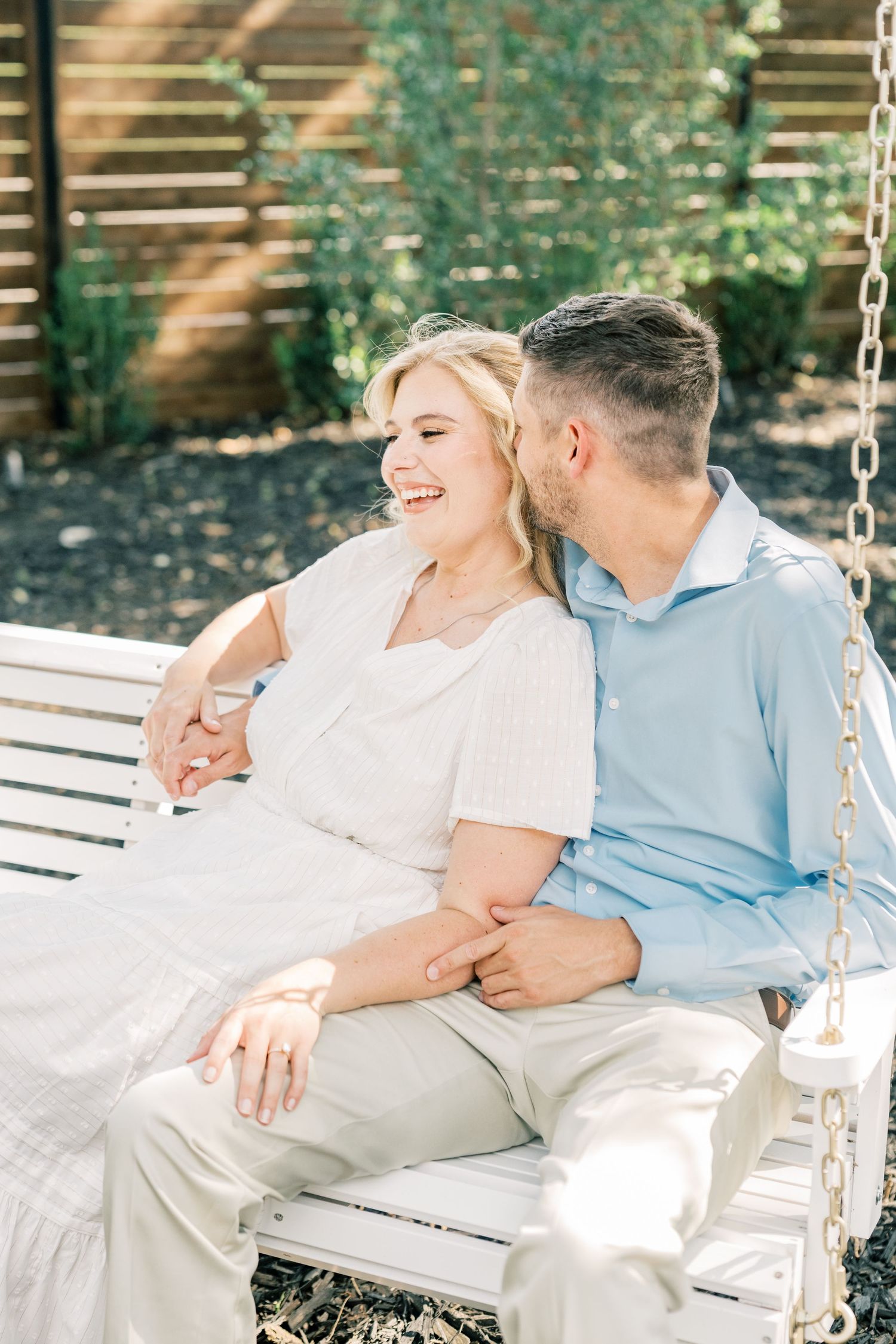 Couple sitting on white swing bench outdoors, man kissing woman's cheek as she laughs joyfully.