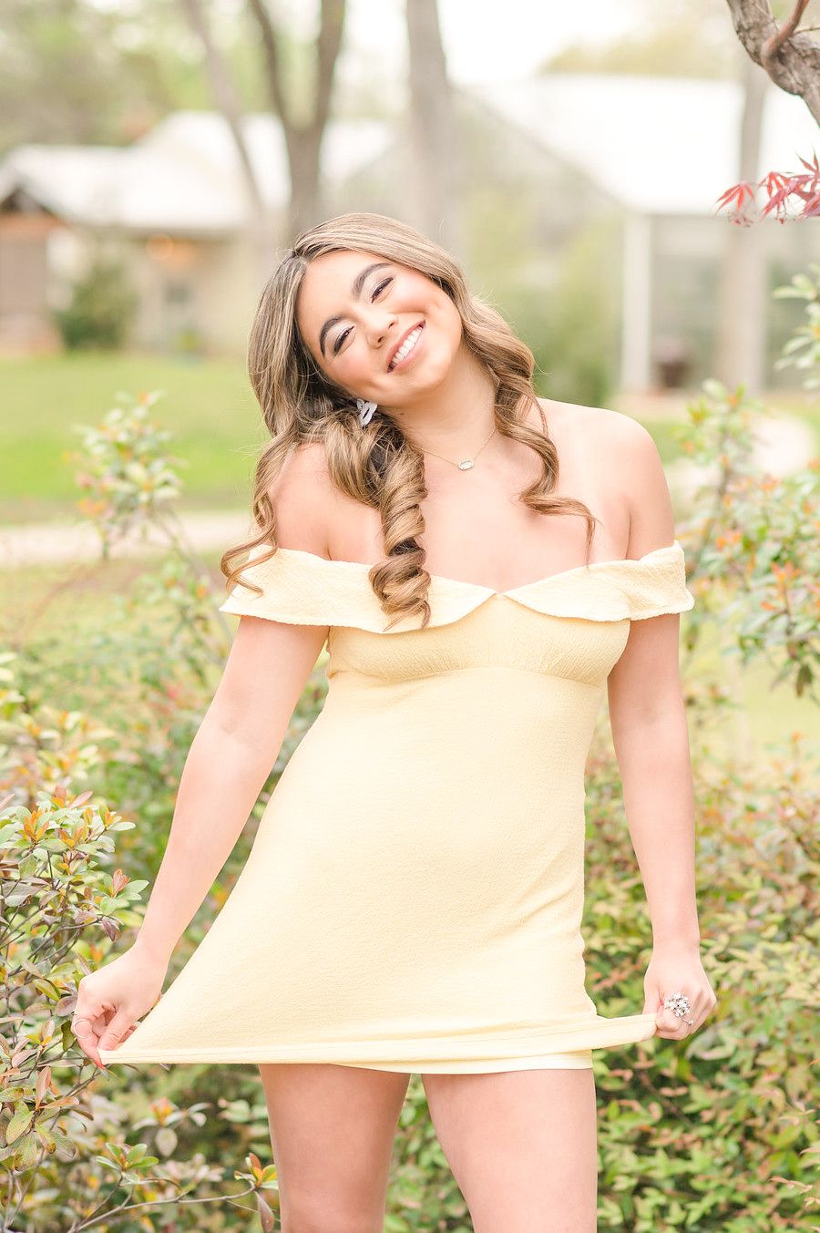 Young woman in yellow off-shoulder dress smiles with braided hair in outdoor garden.