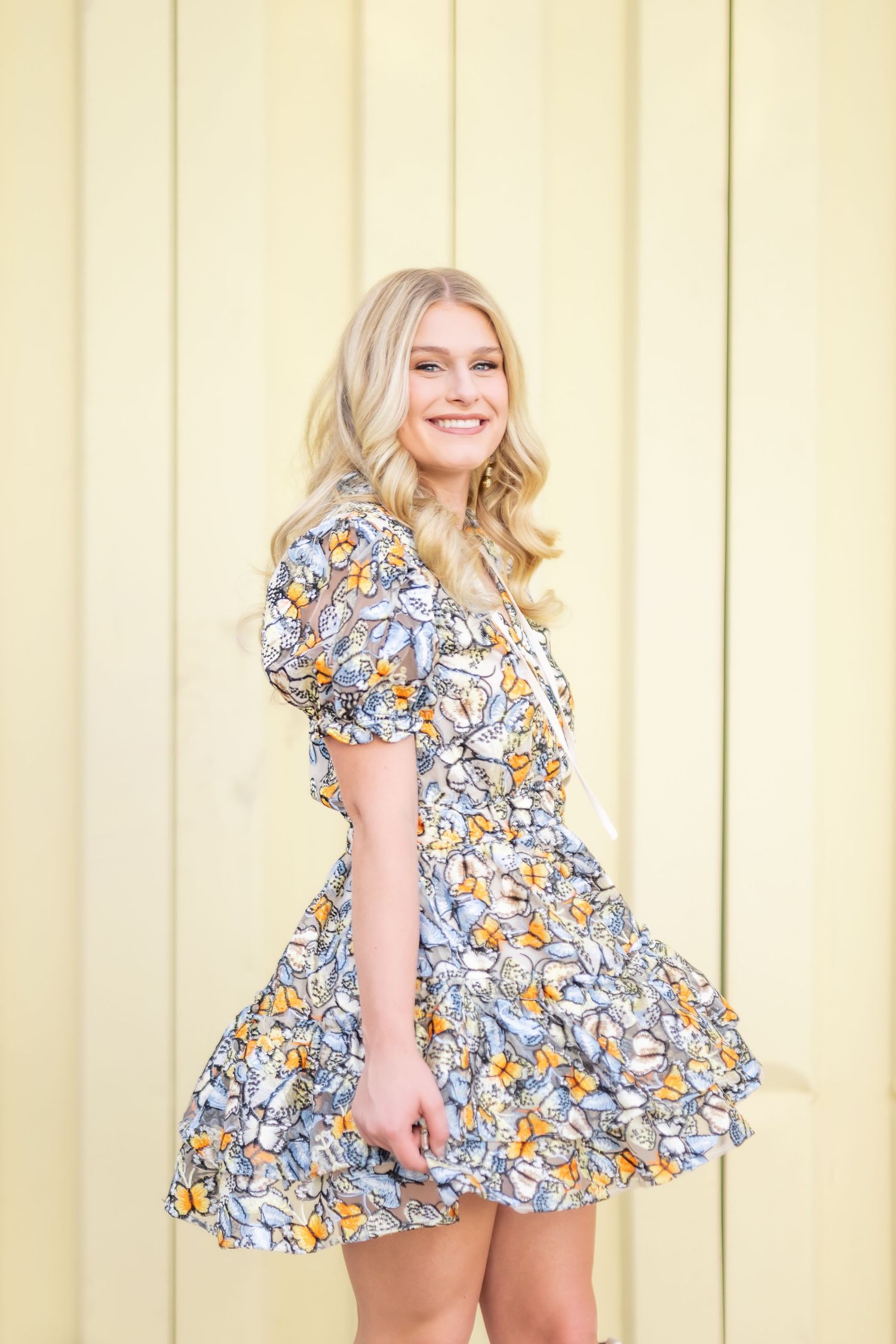 Smiling blonde woman twirling in a floral butterfly print mini dress against a yellow wall.