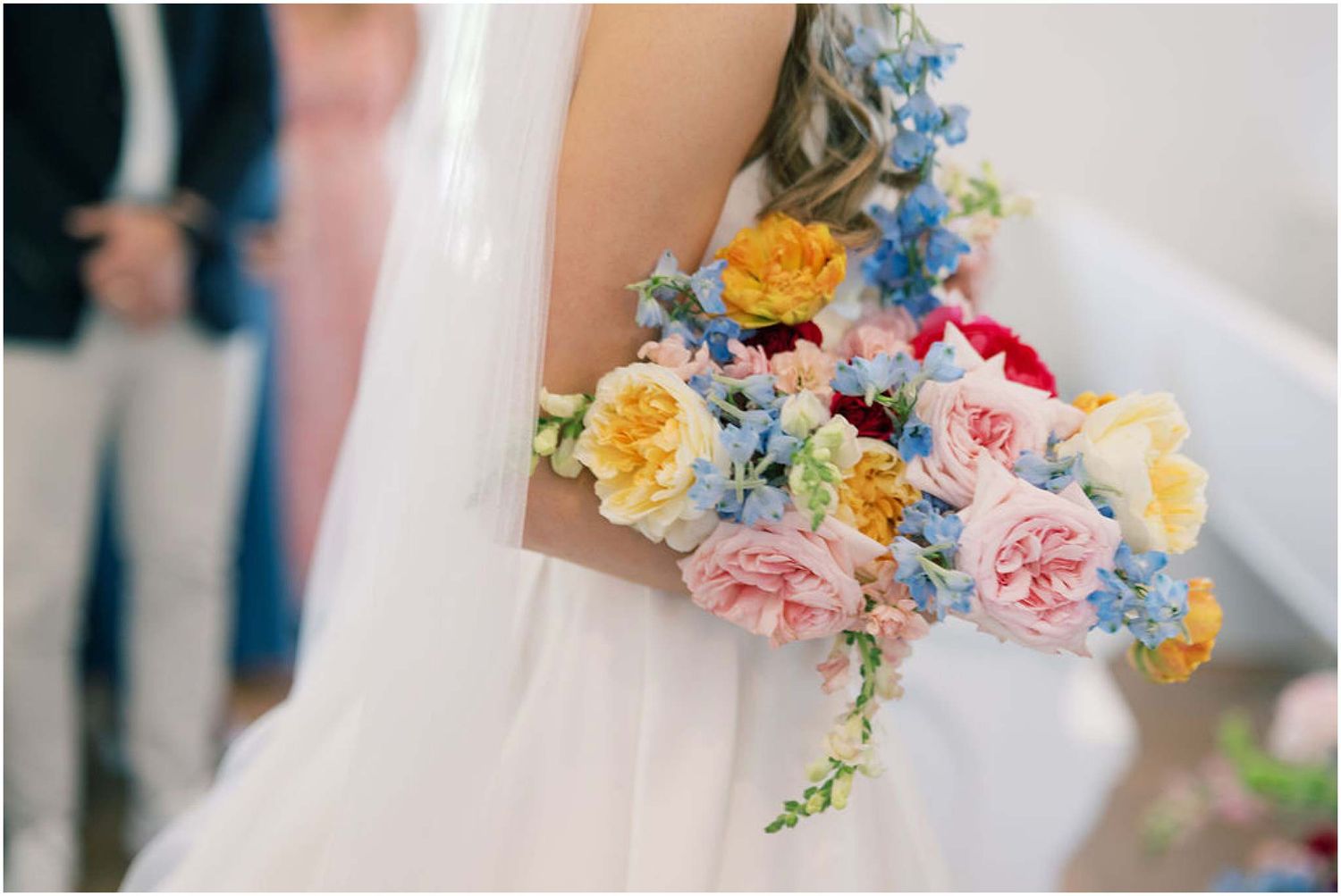 Bride holding a vibrant wedding bouquet with pink, yellow, red, and blue flowers against a white dress.
