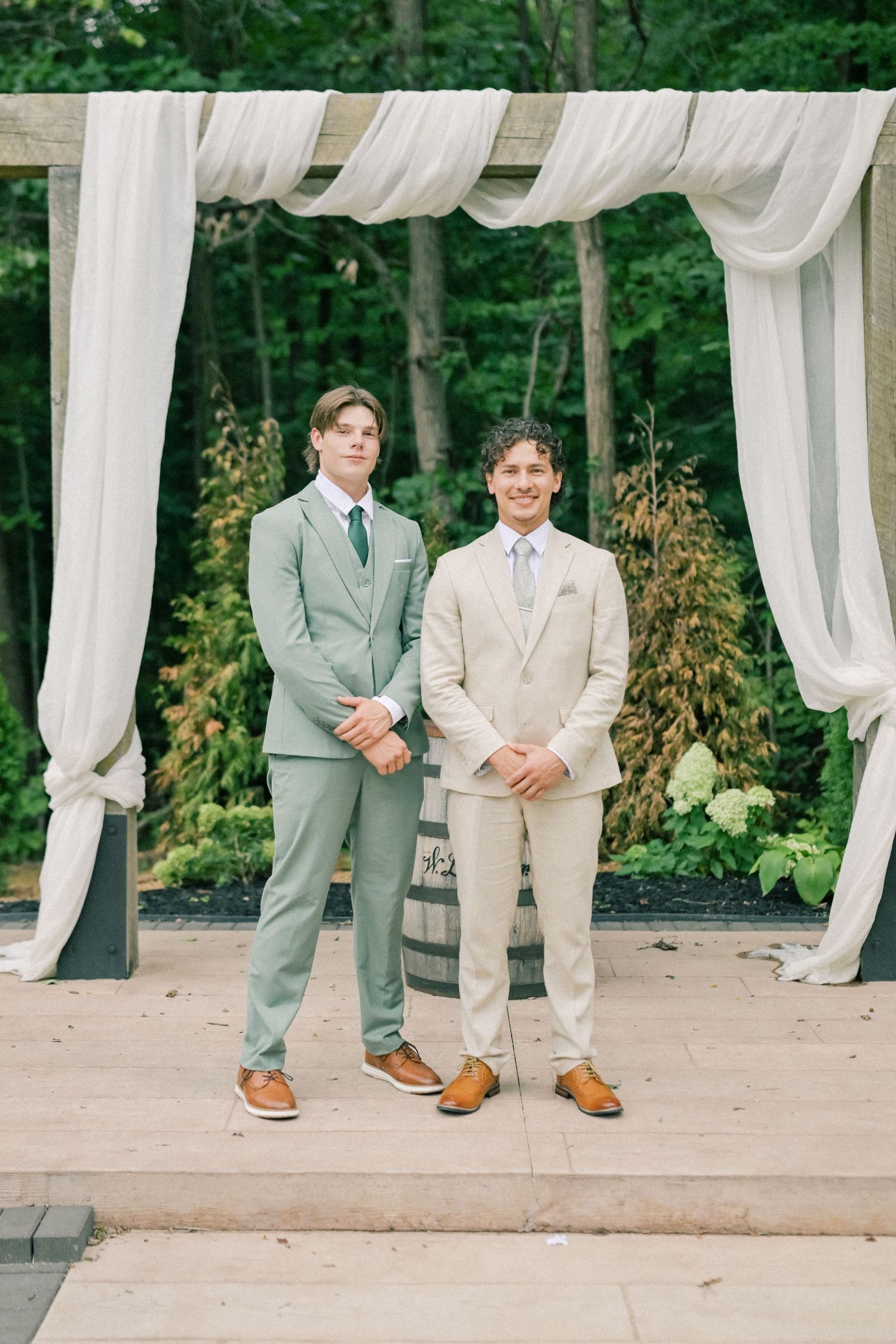 Two grooms in sage green and beige suits pose under a draped white pergola at an outdoor wedding ceremony.