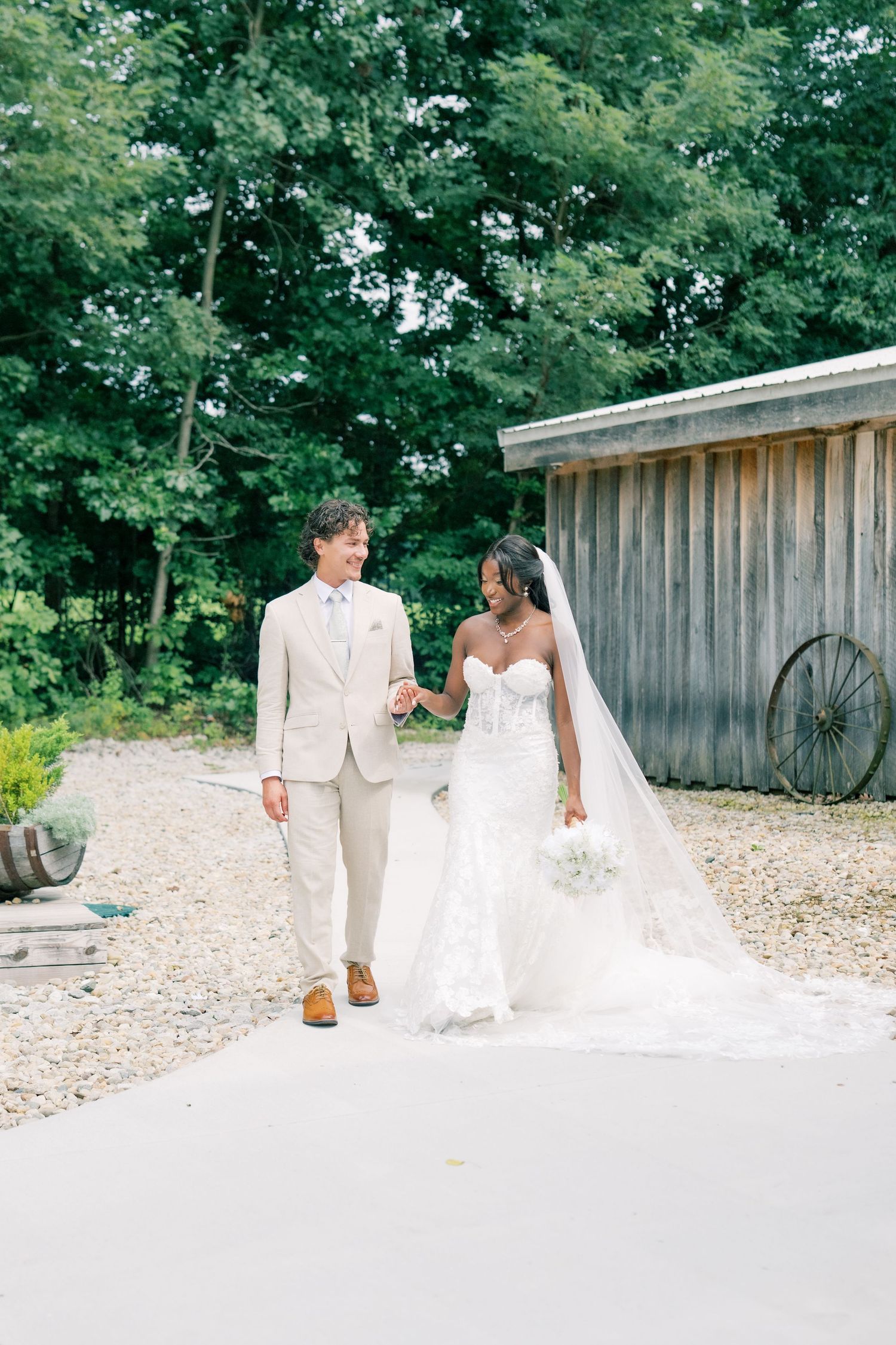 Interracial couple walking hand in hand on wedding day, bride in lace gown with veil, groom in beige suit.