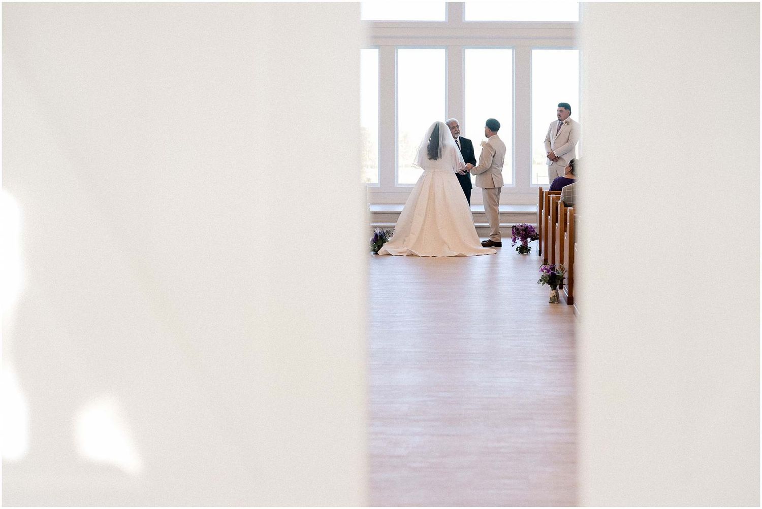 Wedding ceremony guests gather in an elegant white room with high ceilings and natural light.