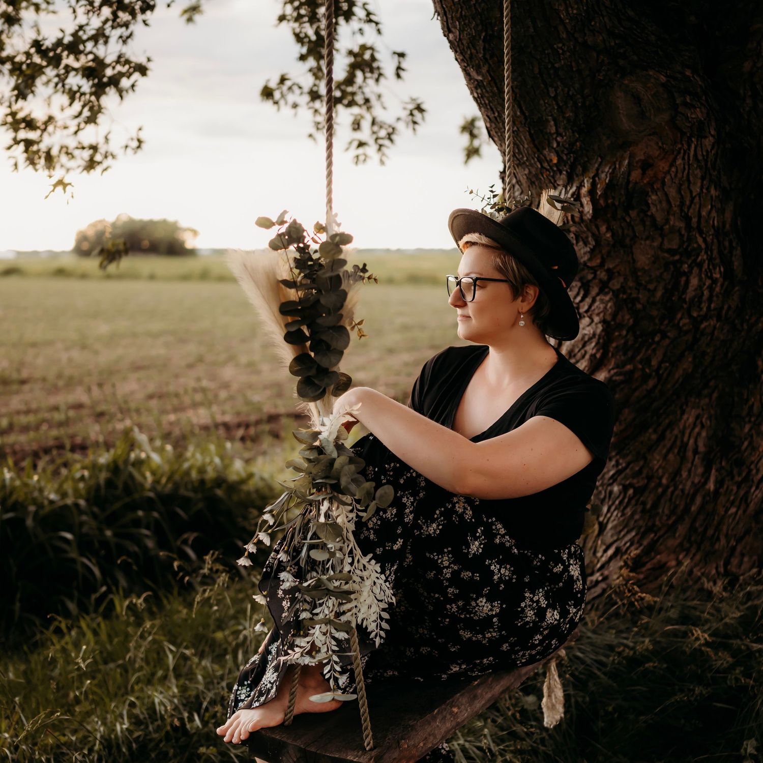 Person in black dress sitting under tree in golden field during sunset, creating dreamy outdoor portraits.