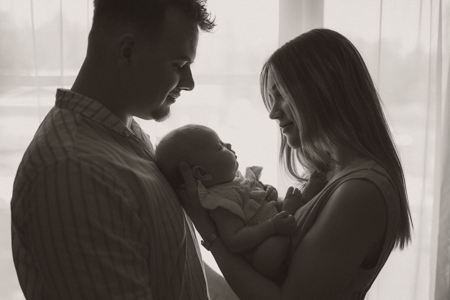 Black and white photo of new parents lovingly gazing at their newborn baby by a bright window.