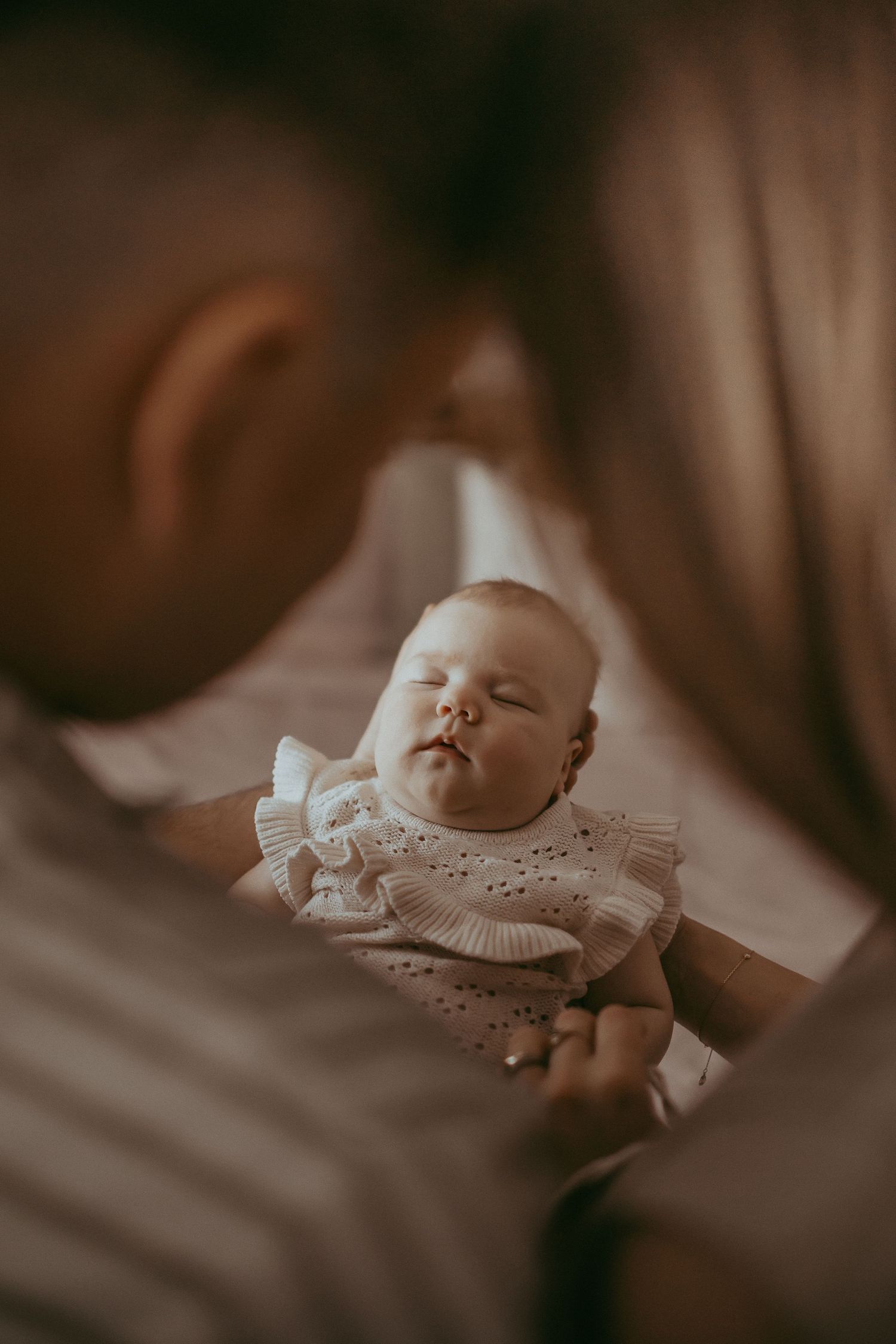 Parents tenderly holding their sleeping newborn baby dressed in white ruffled outfit in a warm intimate moment.