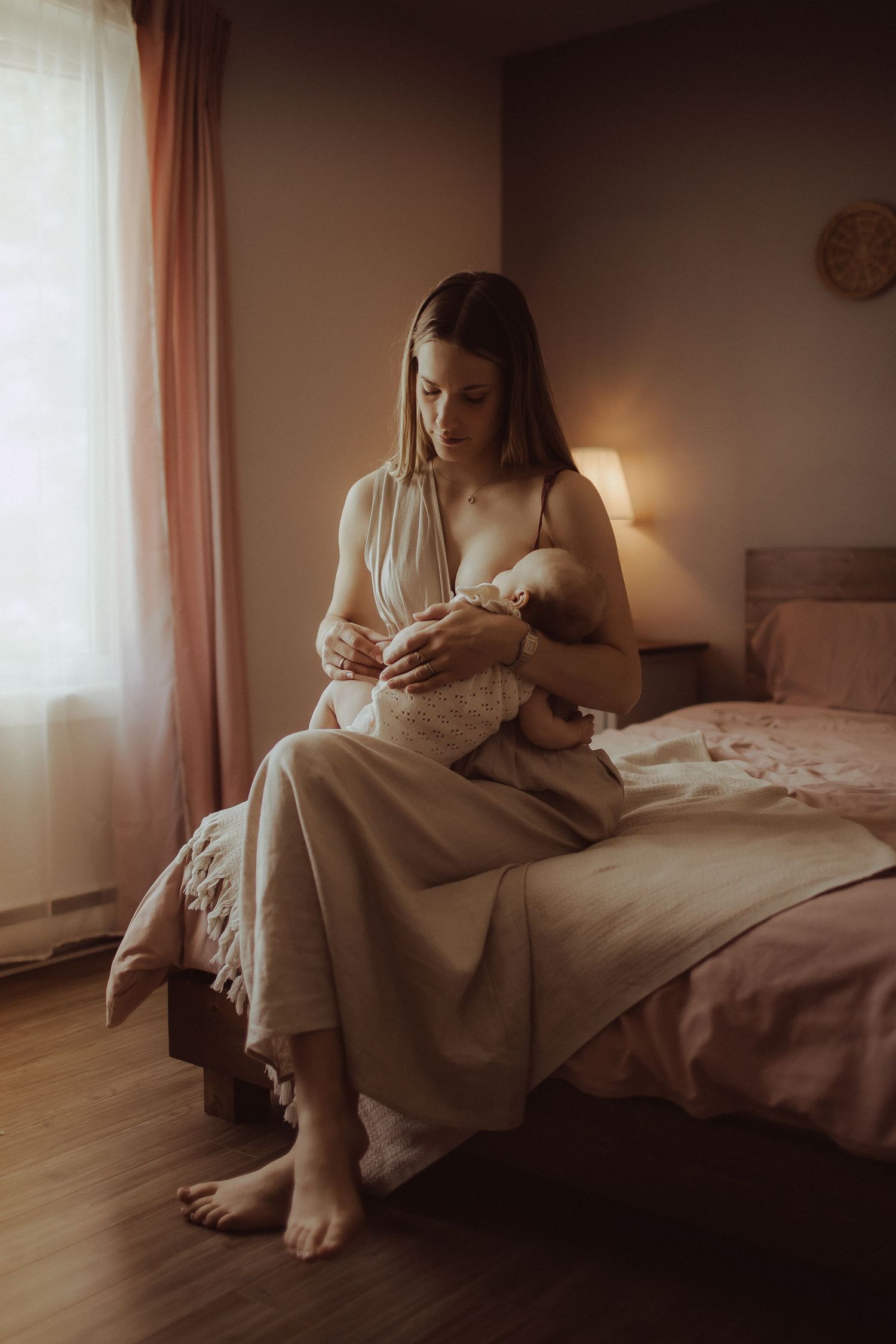 A barefoot mother in a beige dress sits on a bed breastfeeding her newborn baby in a warmly lit bedroom.