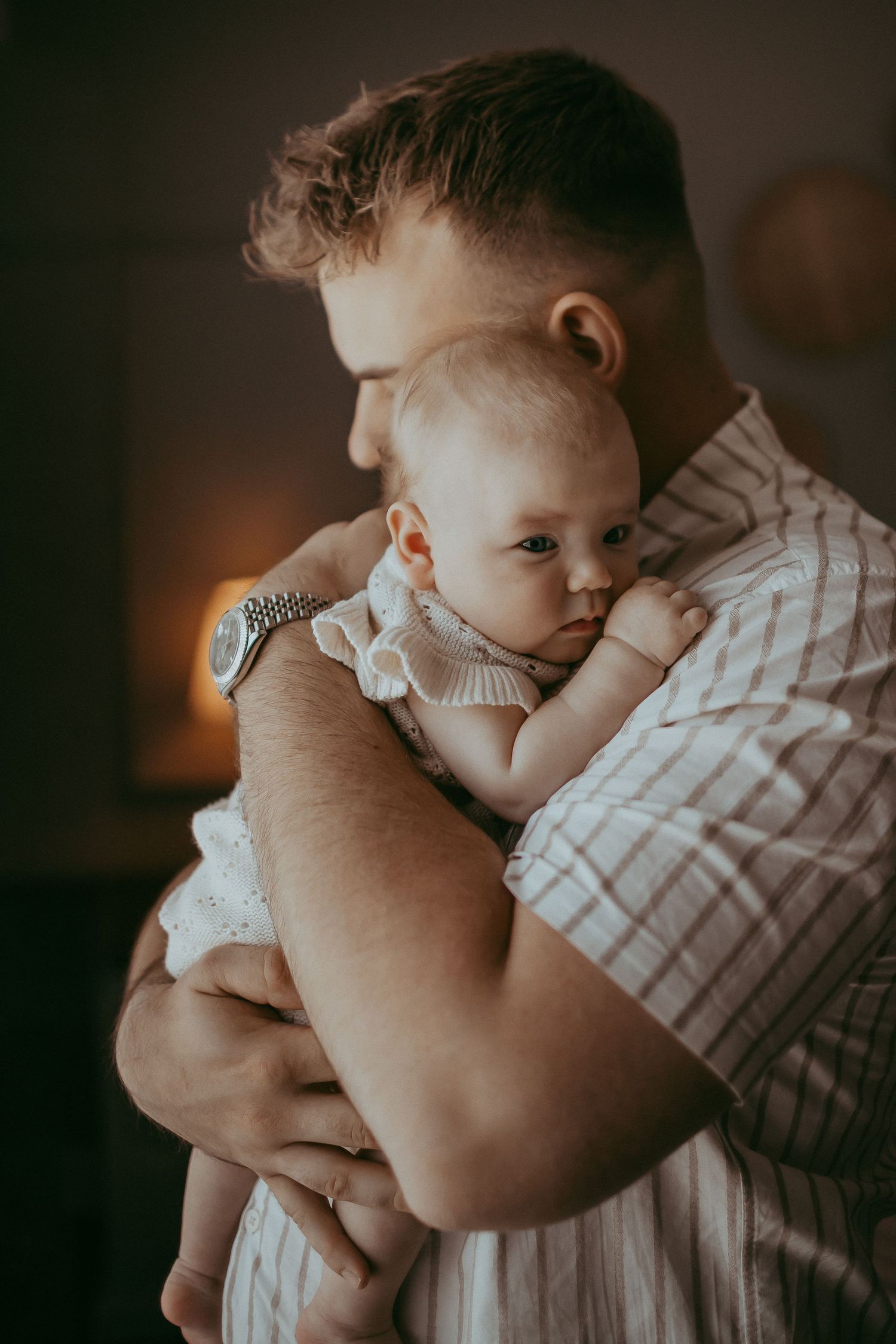 Father tenderly holding newborn baby against his chest in warm, dimly lit room, wearing striped shirt and silver watch.