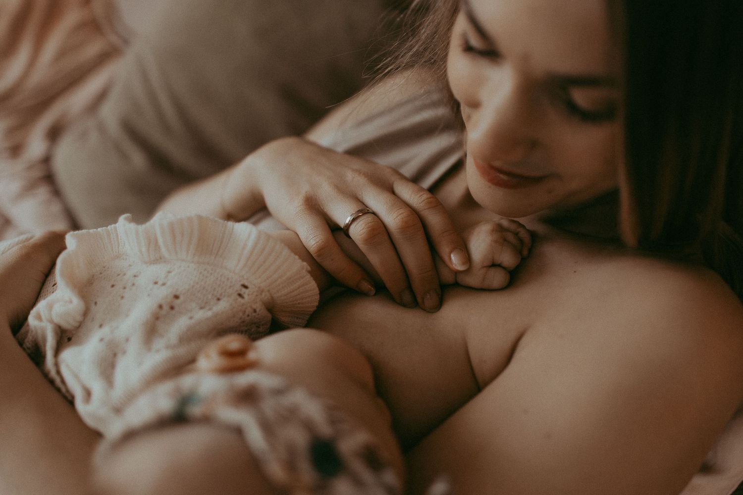 A mother tenderly gazes at her newborn baby while breastfeeding, wearing a wedding ring, in a soft warm-toned setting.