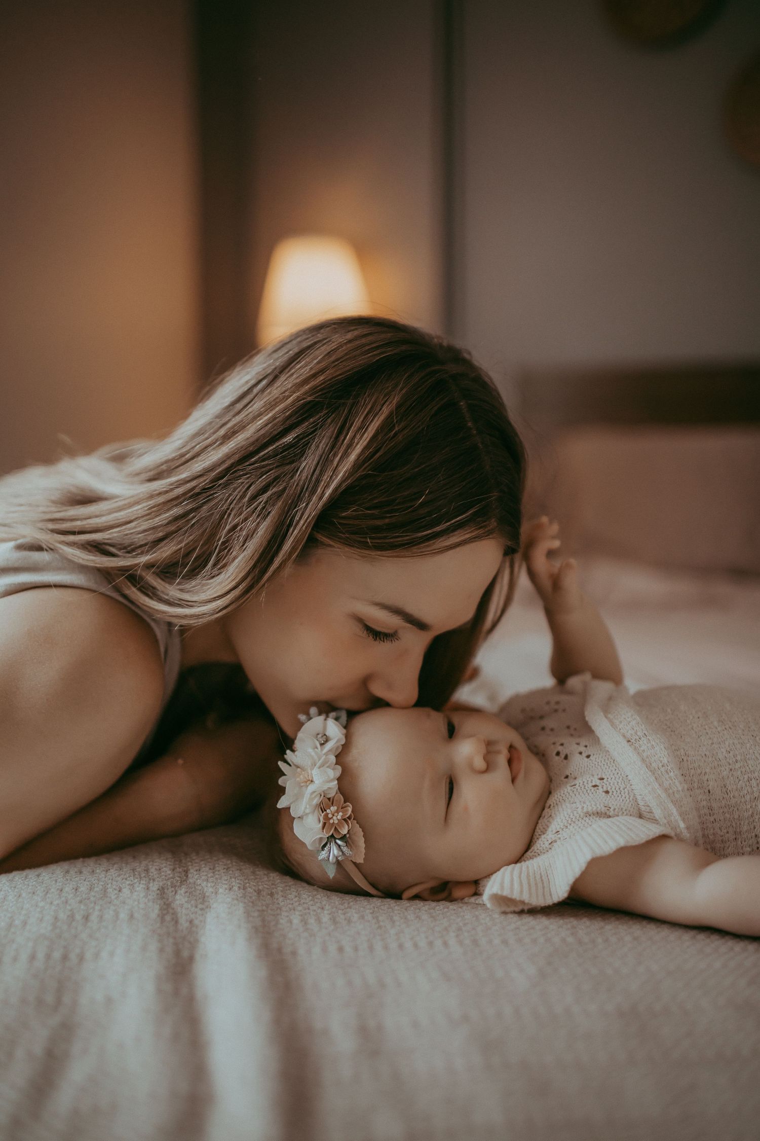 A tender mother gently kisses her newborn baby lying on a bed, with soft warm lighting in the background.