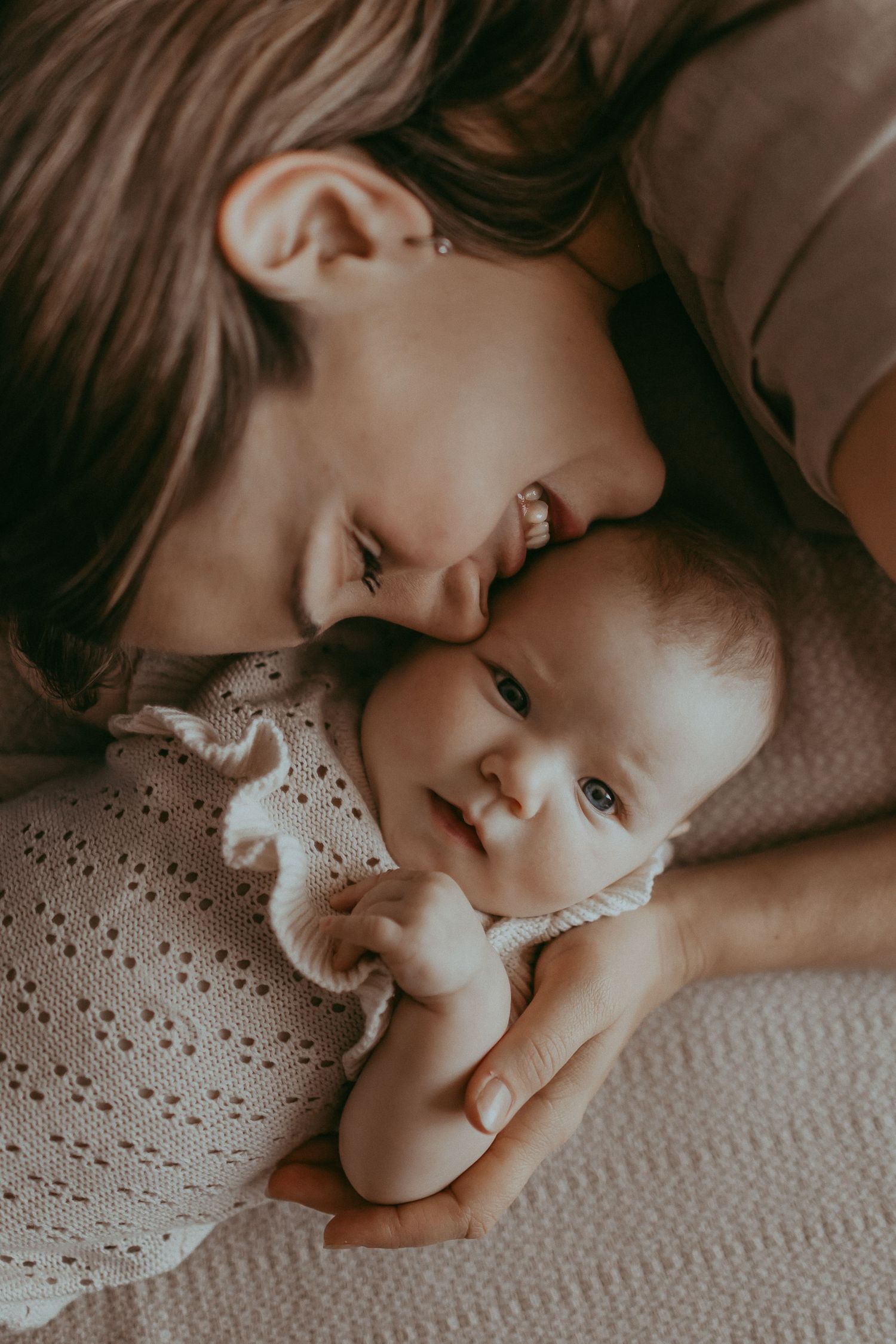 A mother tenderly nuzzles her newborn baby lying on a soft blanket in a warm, intimate portrait.