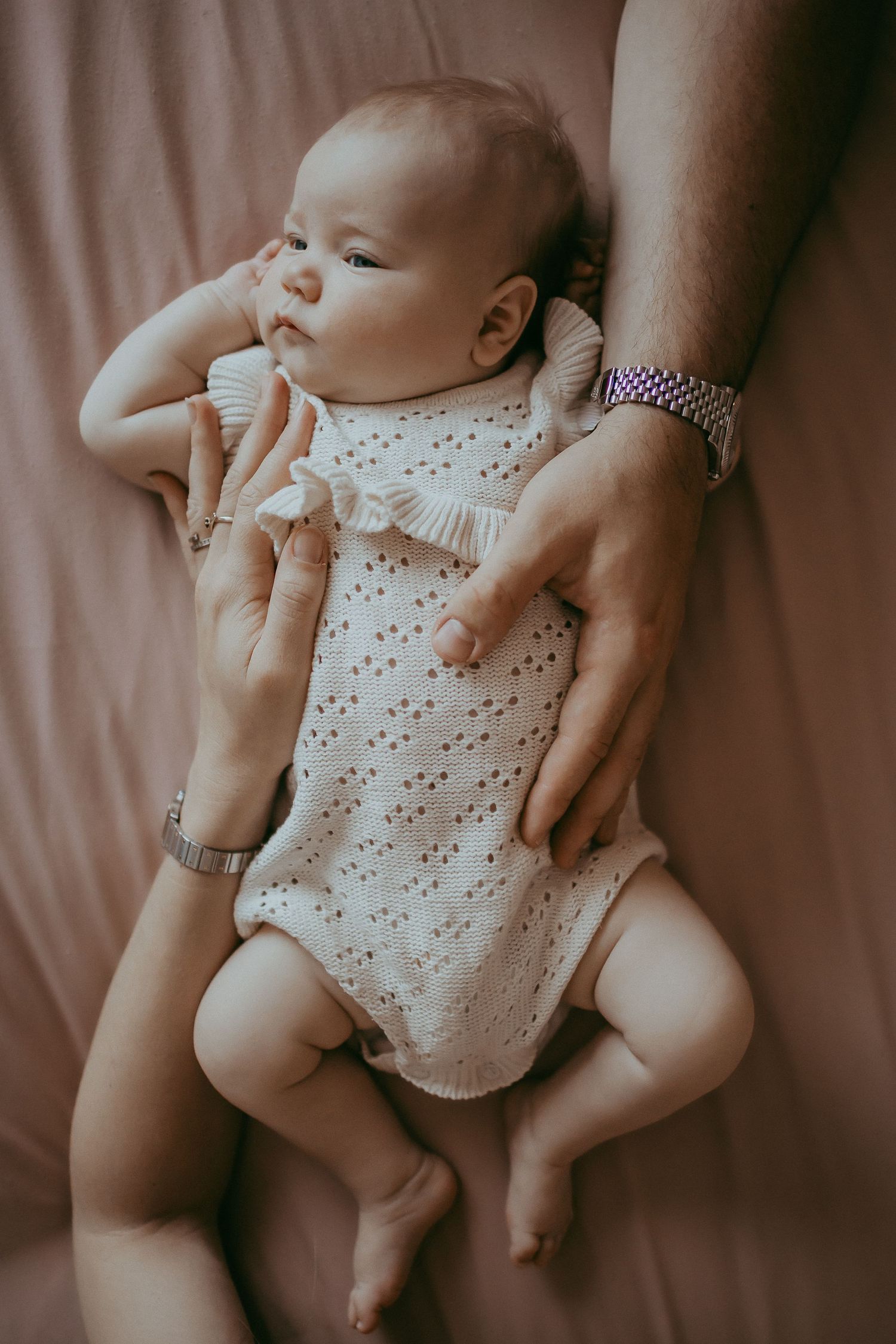 Newborn baby in white knit romper lying on pink blanket, gently held by parents hands wearing watches.
