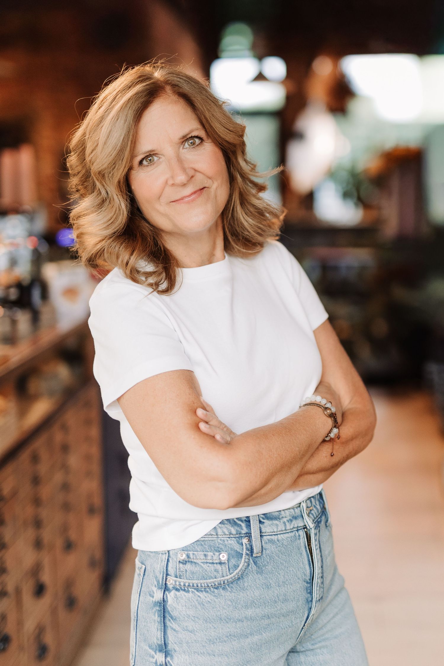 Confident middle-aged woman in white tee and jeans poses arms crossed in cafe.
