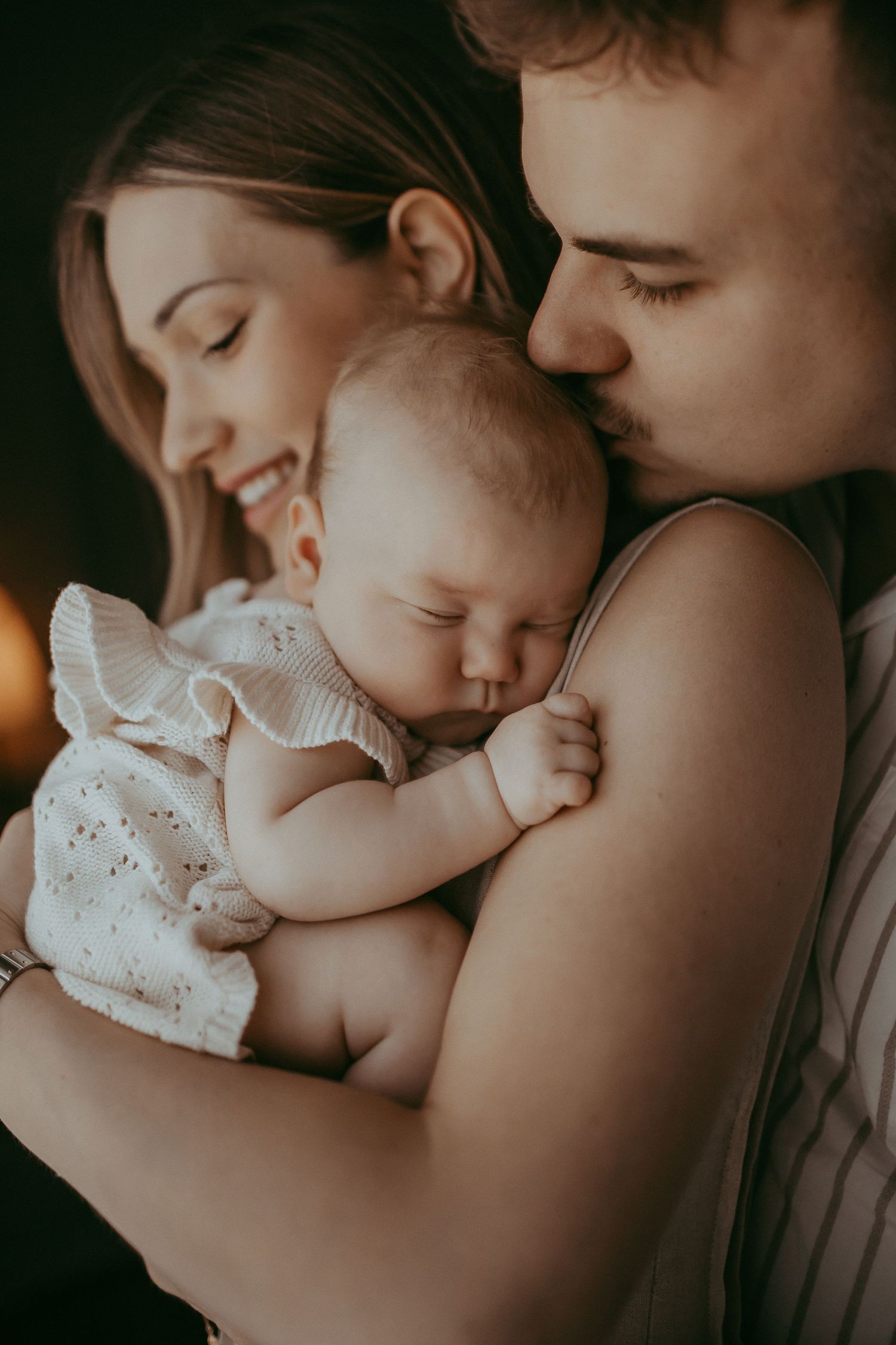 Young parents tenderly holding their sleeping newborn baby, father kissing infant's head in warm intimate family portrait.