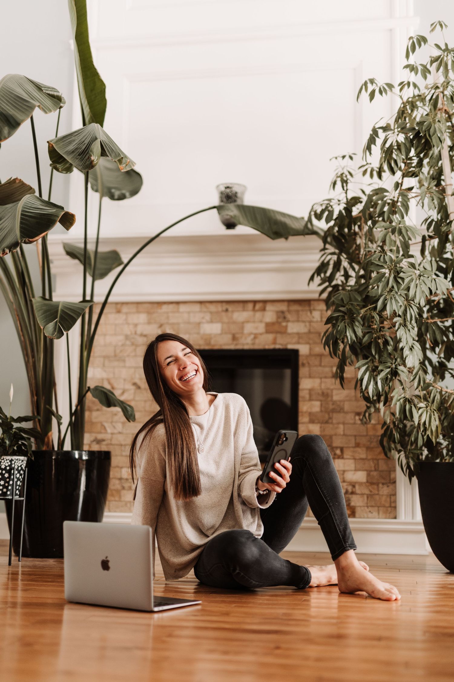 Smiling woman sitting on hardwood floor with MacBook laptop and smartphone, surrounded by lush indoor plants near a brick fireplace.