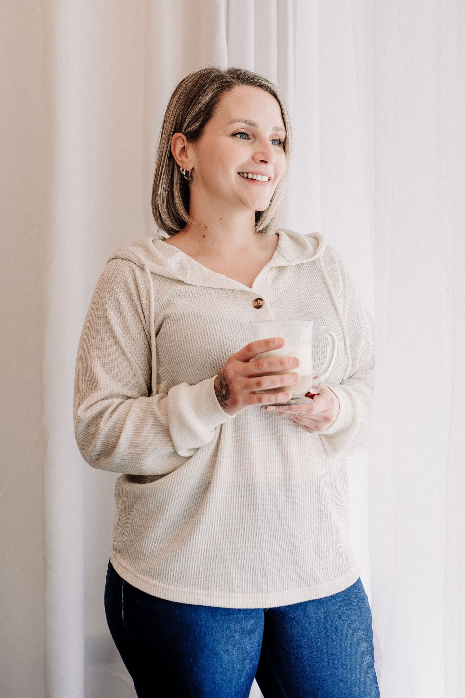 Smiling woman in beige hoodie holding white mug, standing by bright window in blue jeans.