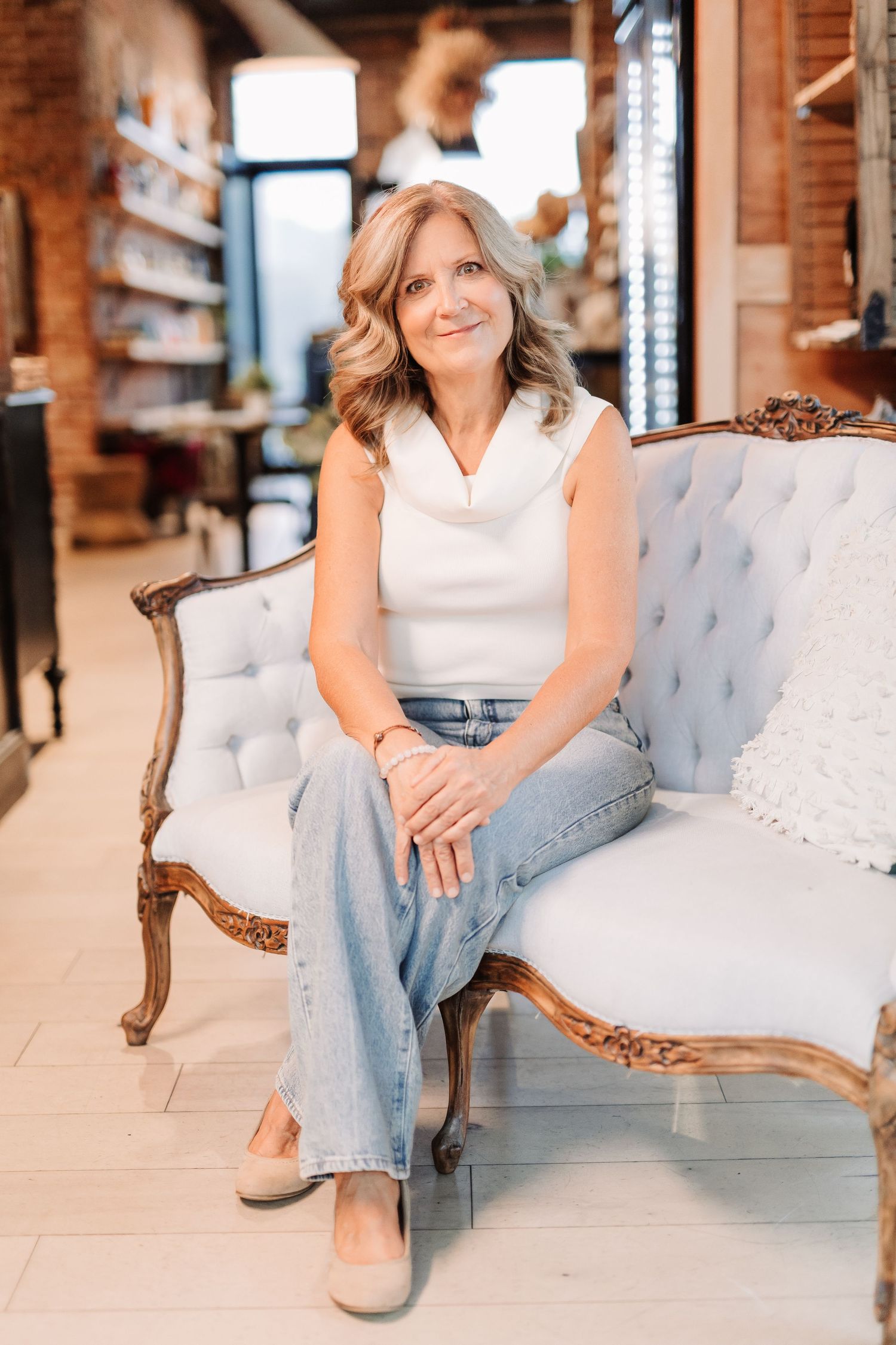 Smiling woman in white top and jeans sitting on vintage tufted sofa in a boutique studio.