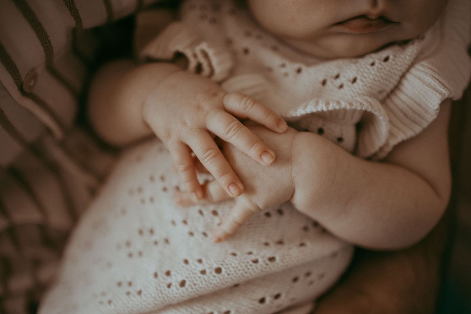 Close-up of a sleeping newborn baby in a white knit outfit, with tiny delicate hands resting gently together.