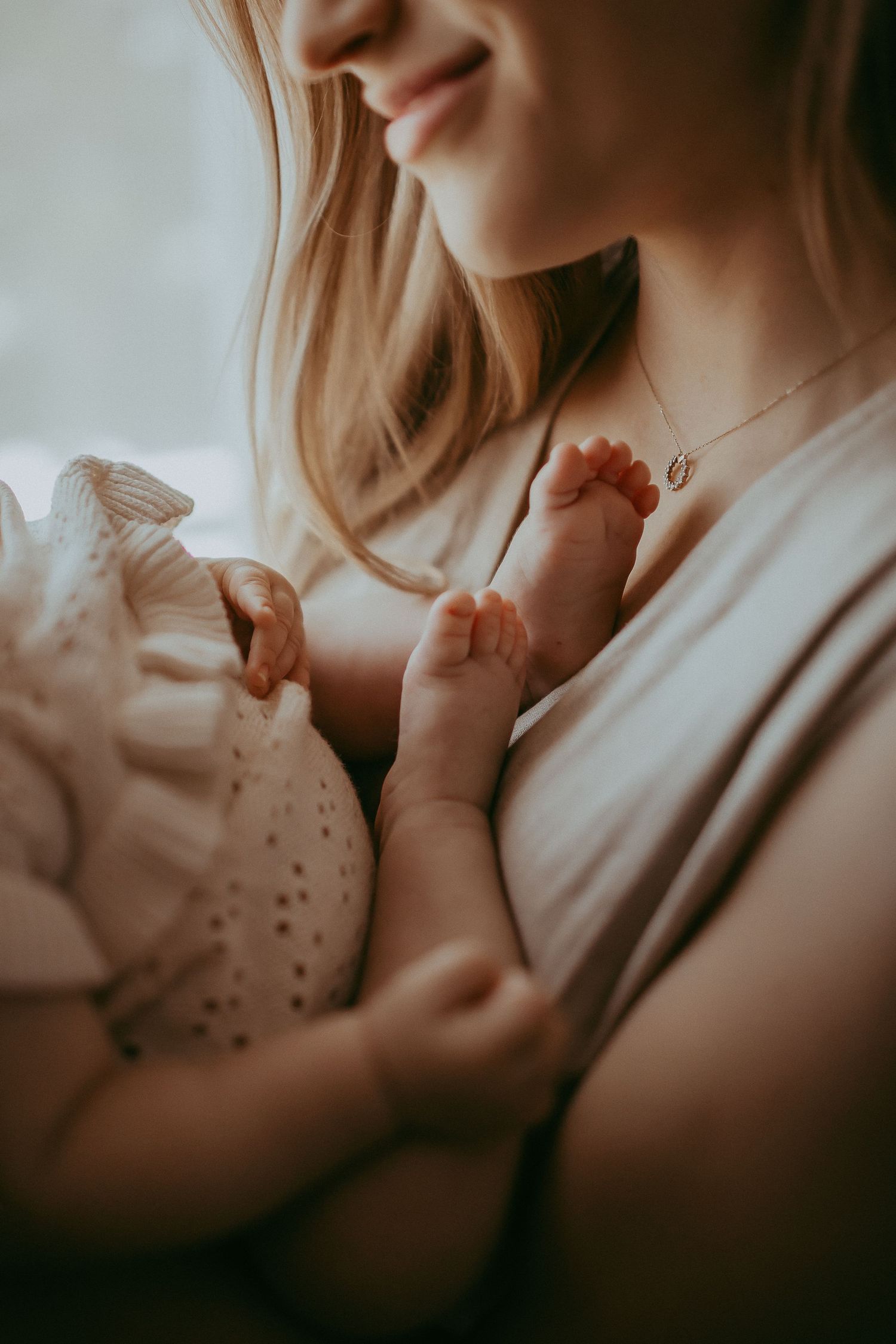 A mother smiling while breastfeeding her newborn baby, wearing a delicate gold necklace.