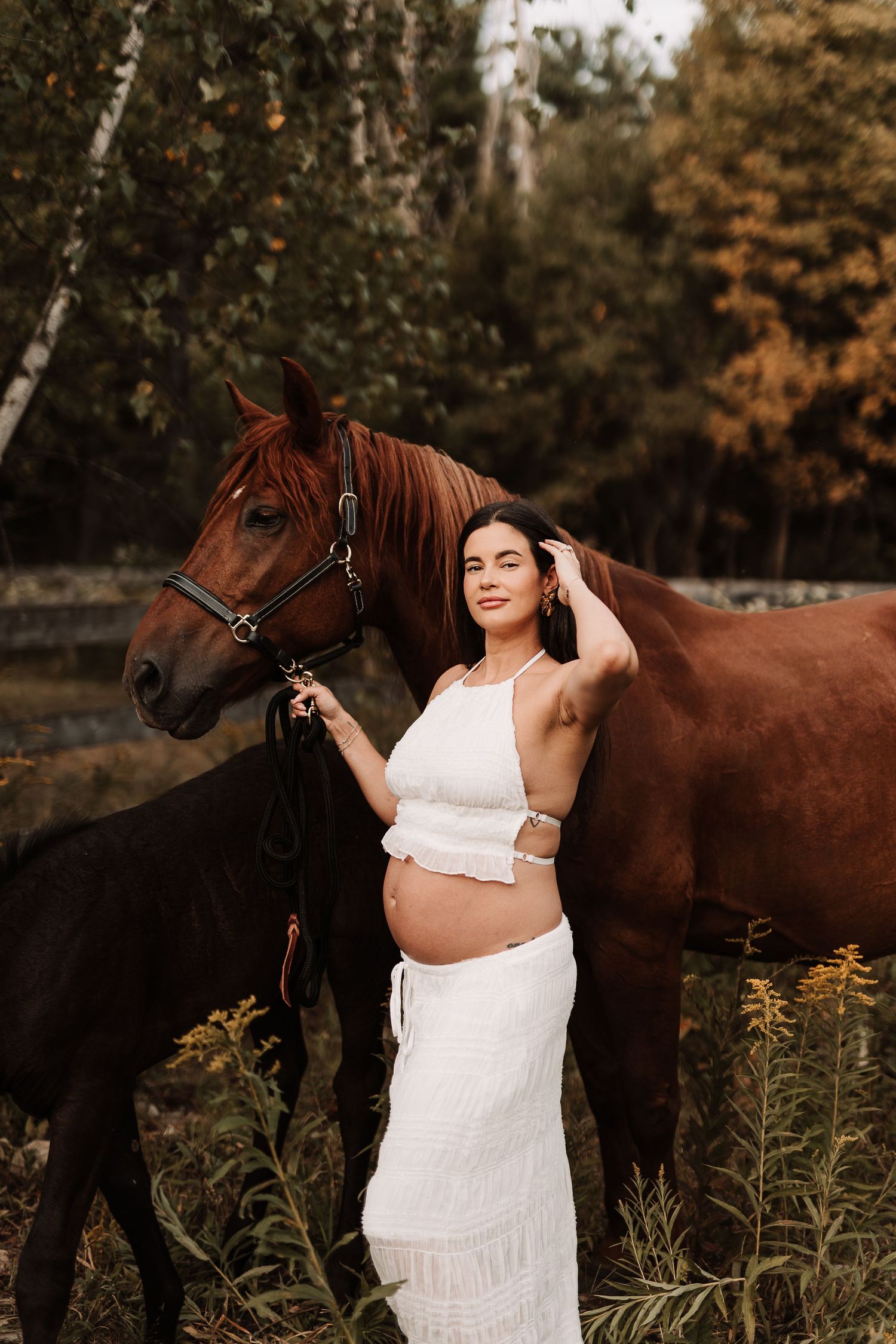 Pregnant photographe, Maternité, champ, rive-sud,  in white crop top and skirt stands with chestnut horse and foal in autumn forest setting.