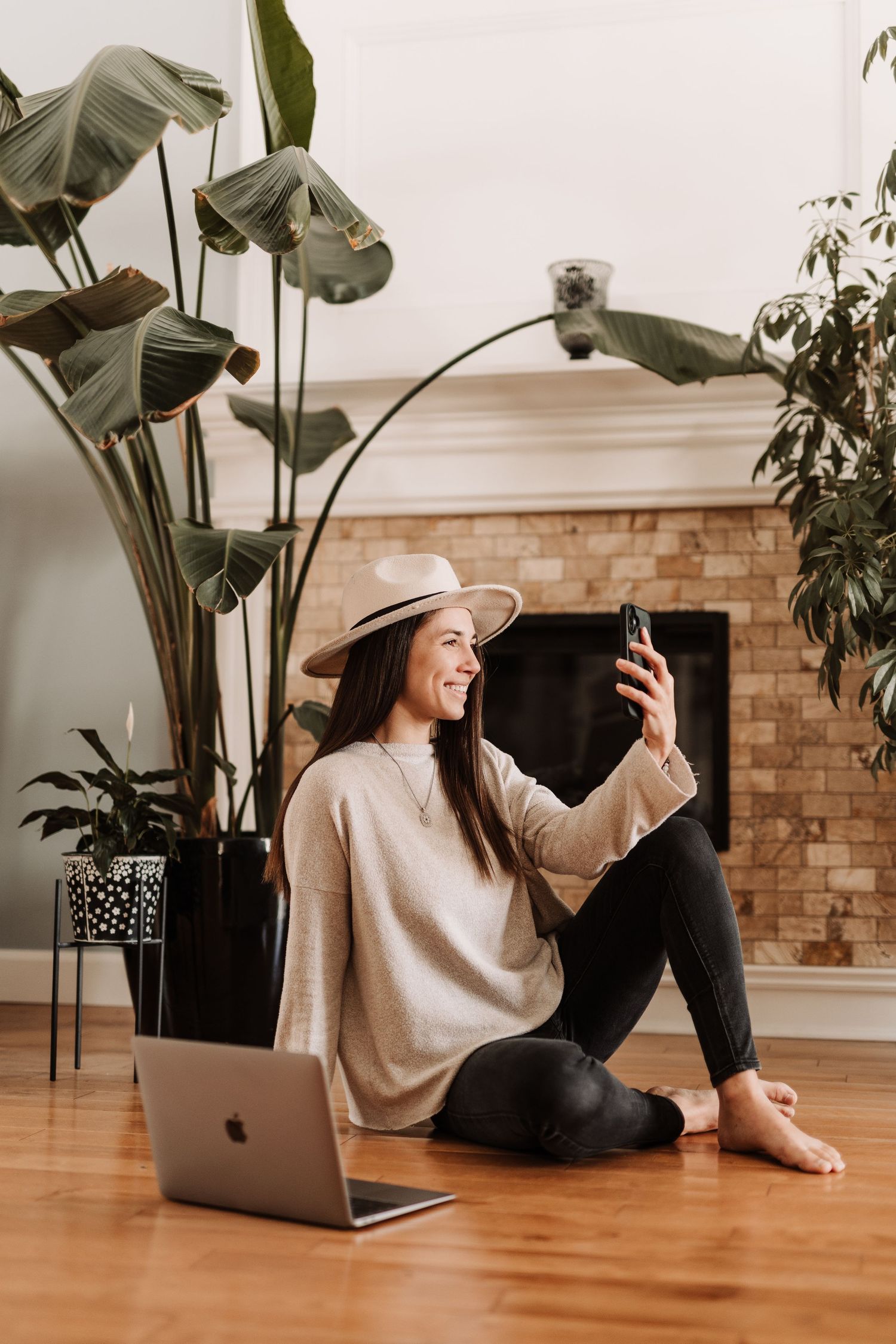 Woman in beige hat and sweater sitting on hardwood floor, taking selfie with phone near laptop and large tropical plant.