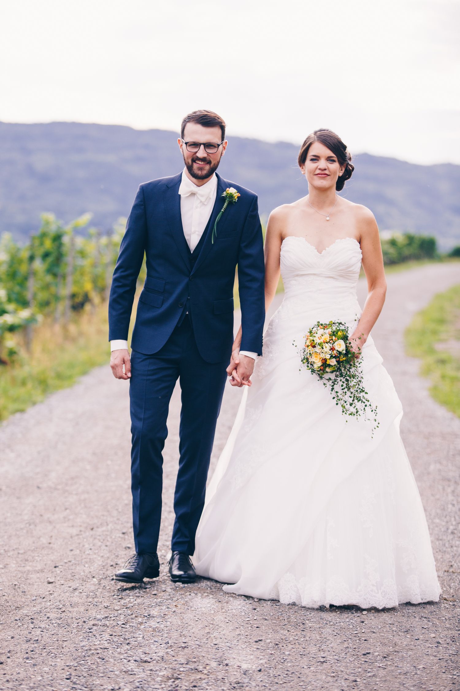 Bride and groom holding hands walking on a rural path, she in white lace gown with bouquet, he in navy suit.