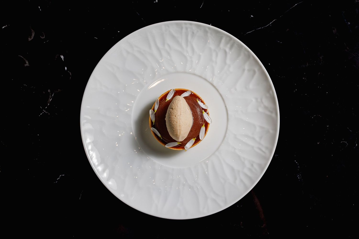 Elegant dessert with a smooth dome and caramel sauce on a white embossed plate against a dark background.