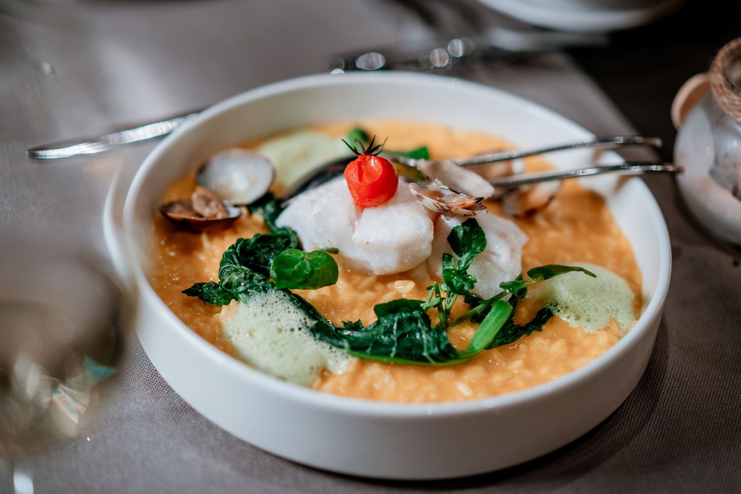 Elegant white bowl with poached fish, sautéed greens, cucumber, and creamy tomato bisque garnished with a physalis.