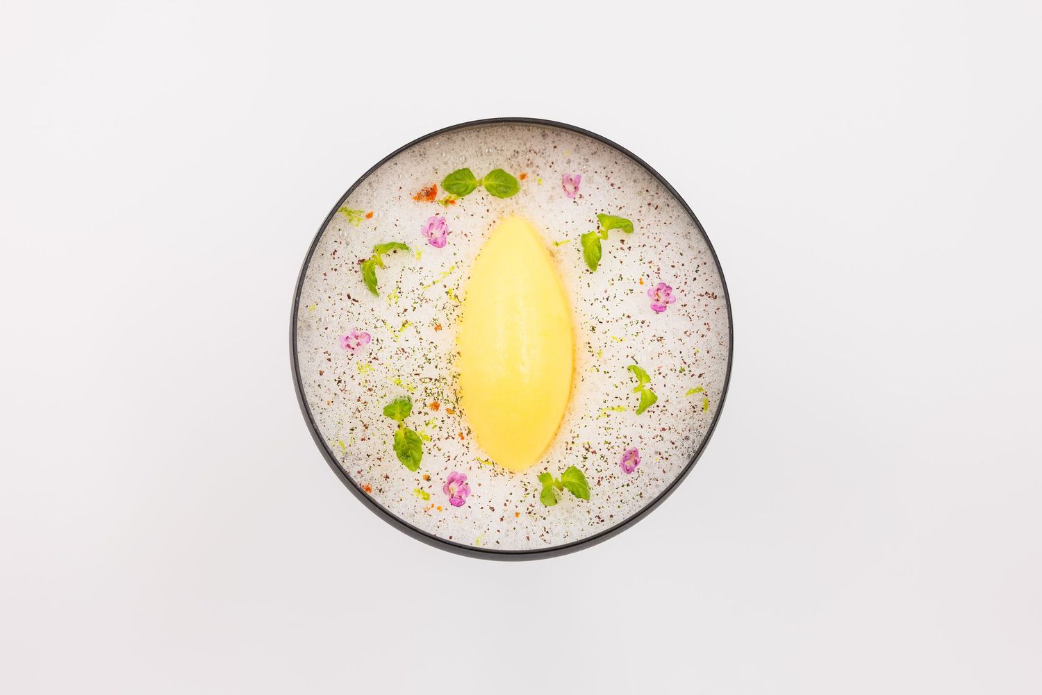Elegant dessert bowl with yellow mango sorbet, edible flowers, and chia seeds on white background.