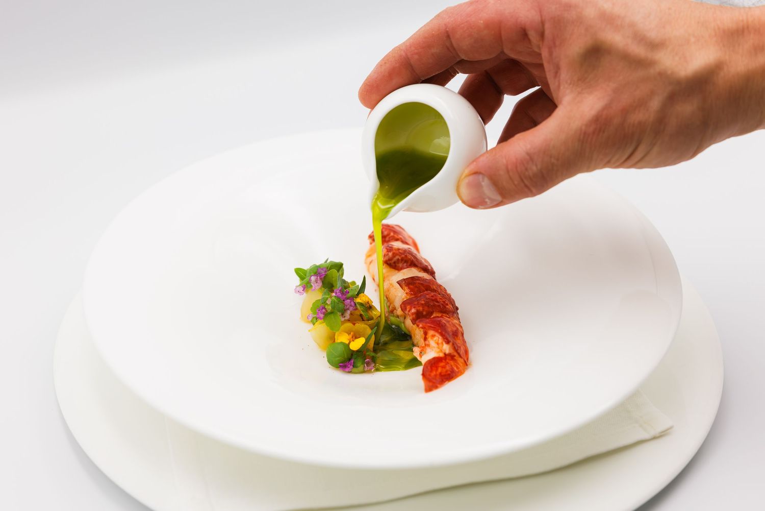 Chef pouring green herb sauce over a plated lobster dish on a white plate in a fine dining presentation.