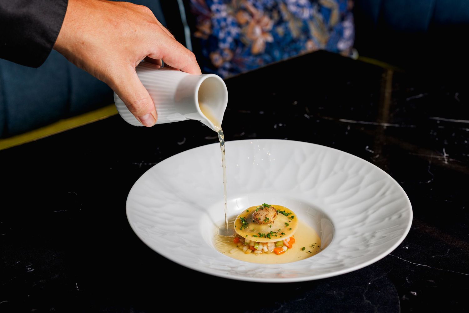 Chef pouring sauce over an elegant gourmet dish on a white plate at a fine dining restaurant.