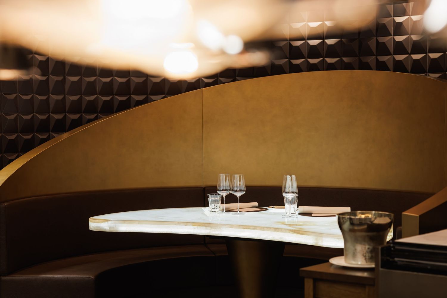 Elegant restaurant booth with curved dark walls, white marble table, and crystal wine glasses in warm ambient lighting.