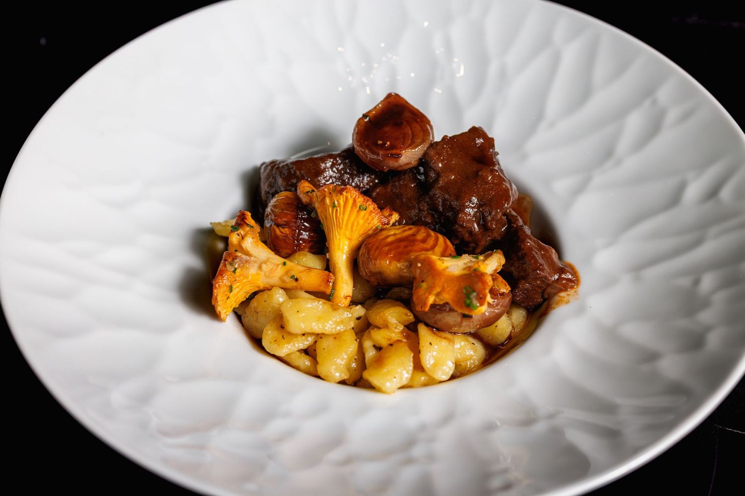 Braised beef short rib with golden chanterelle mushrooms and gnocchi served in a white embossed bowl.