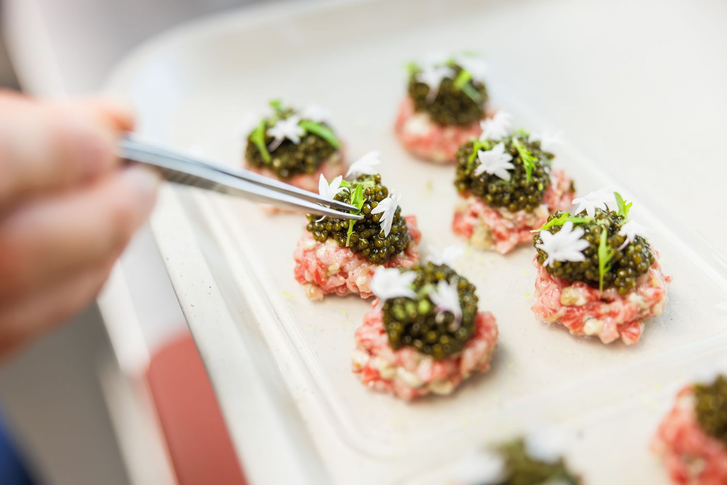 Chef using tweezers to plate gourmet meat tartare bites topped with microgreens on a white serving tray.