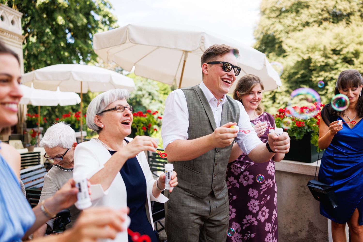 Joyful wedding guests laughing and celebrating outdoors, holding drinks near floral decorations and white umbrellas.