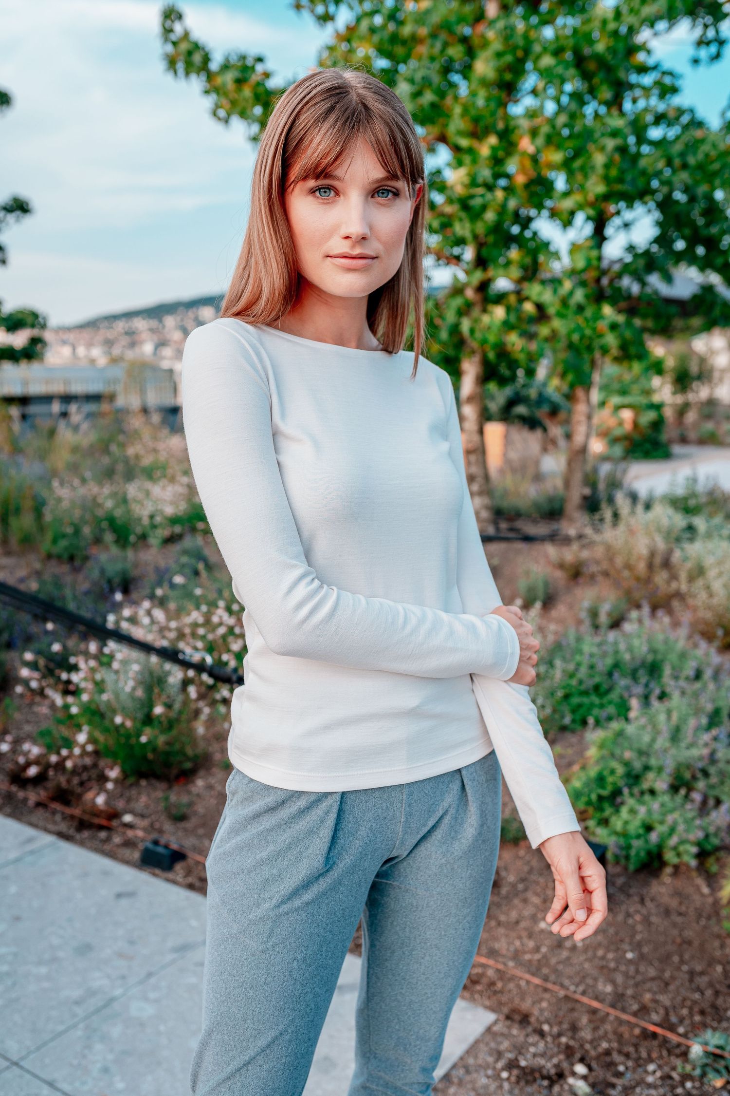 Young woman in white long-sleeve top and gray pants standing outdoors in a garden setting.