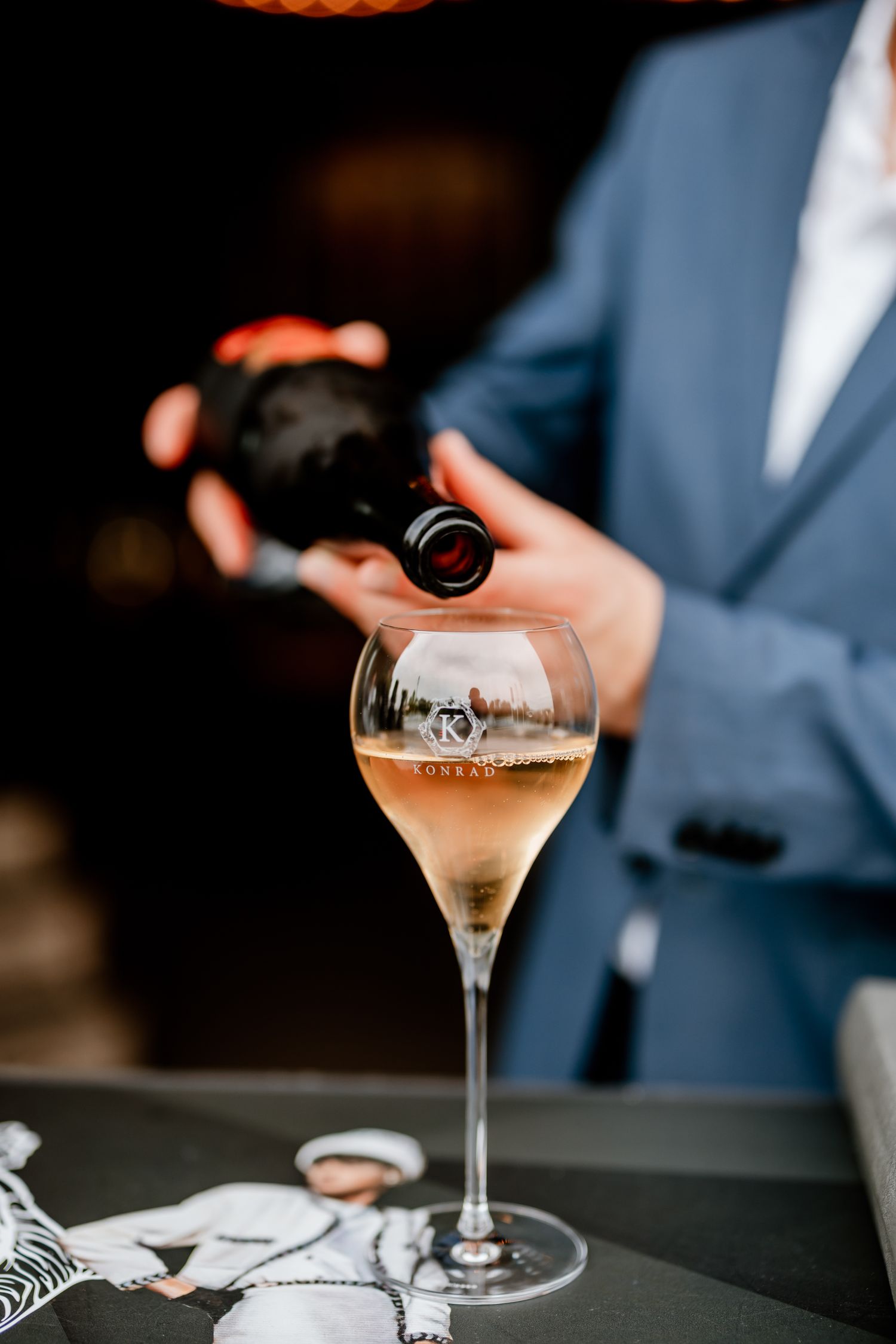 A sommelier in a blue suit pouring rosé wine into an elegant stemmed glass etched with a logo.