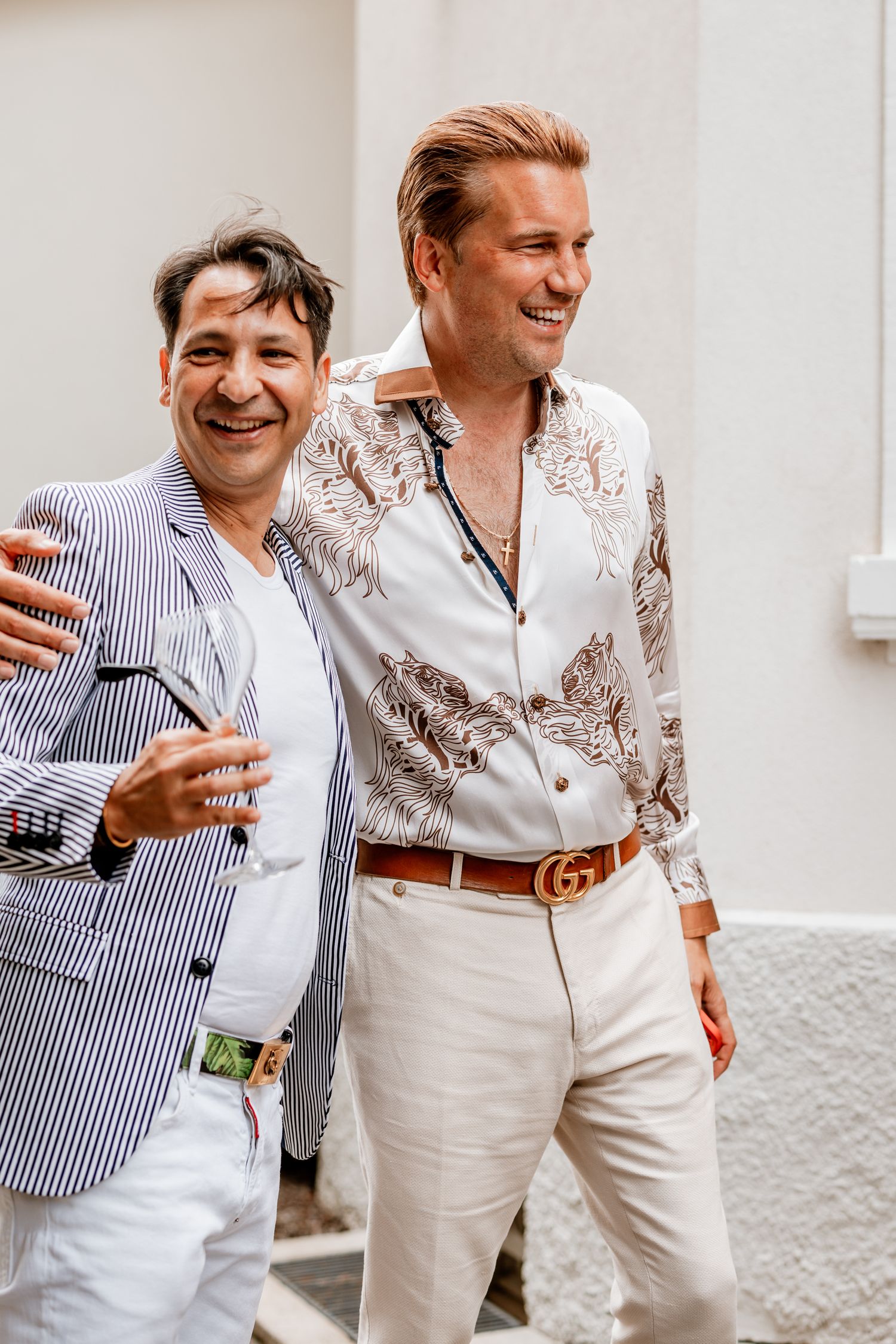 Two stylish men laughing and walking together outdoors, one wearing a Gucci belt and floral shirt.