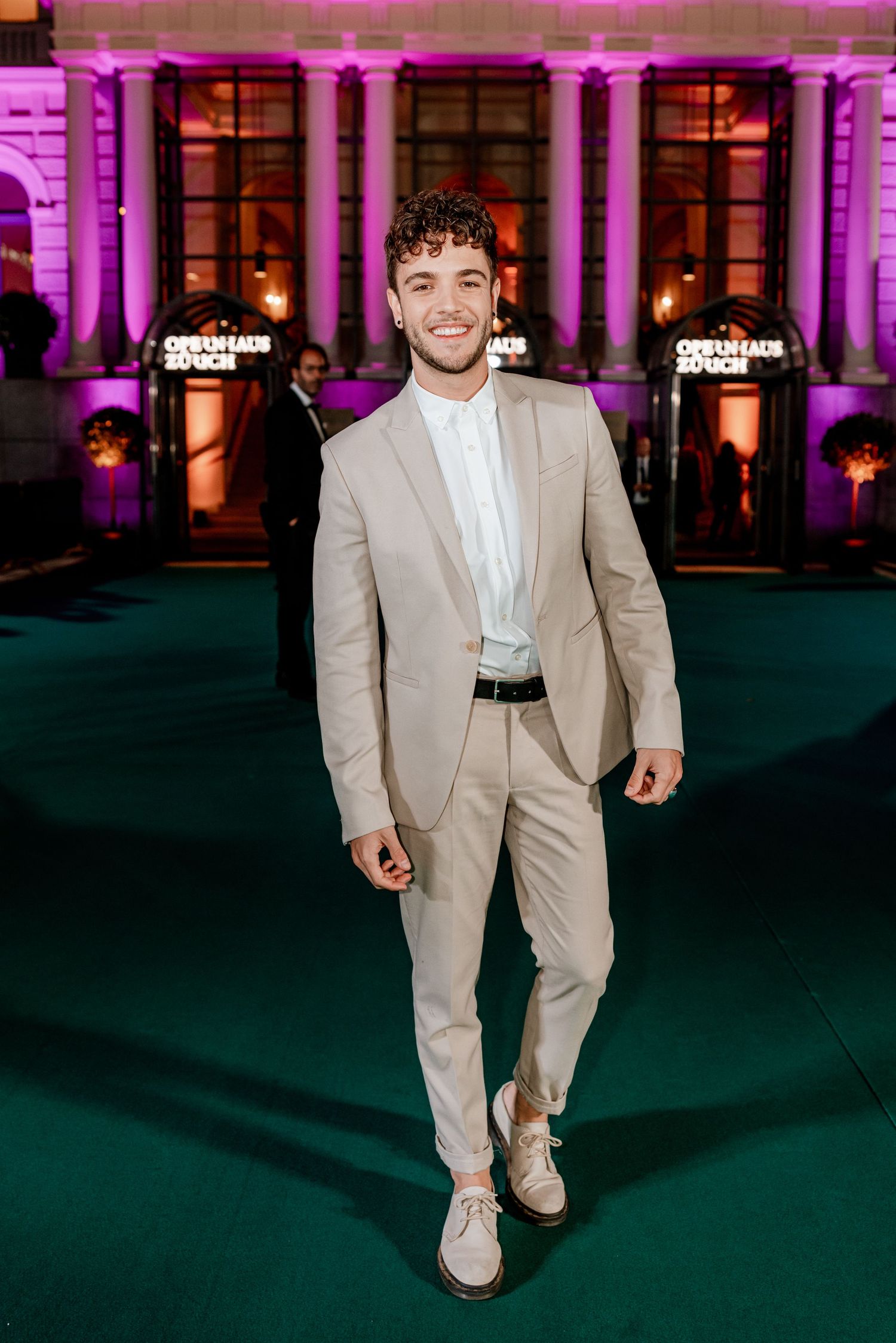 Young man in beige suit poses on green carpet outside illuminated Opernhaus Zurich at night.
