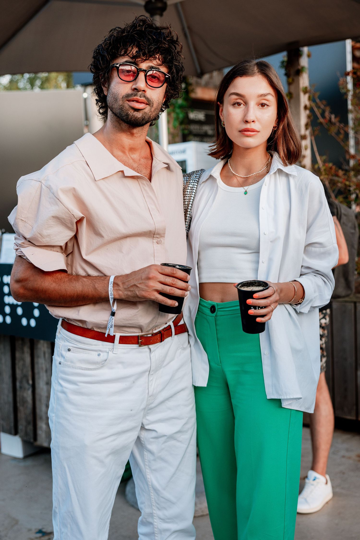 Stylish couple holding coffee cups outdoors, man in beige shirt with sunglasses, woman in green pants and white top.
