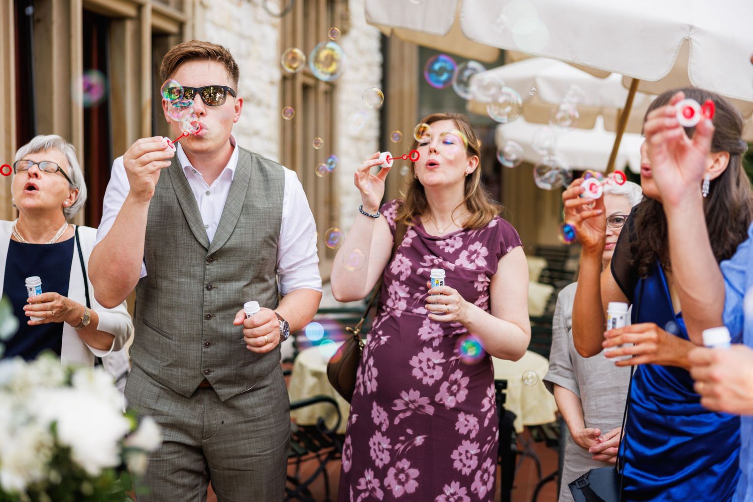 Group of guests blowing bubbles at an outdoor wedding reception, dressed in elegant attire.