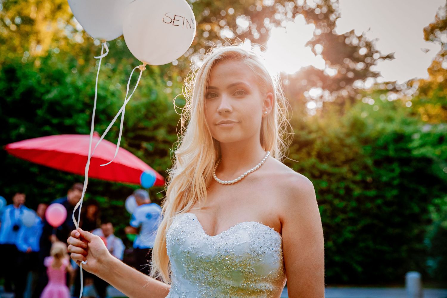 Blonde woman in strapless white gown holding balloons outdoors in golden sunlight at an elegant event.