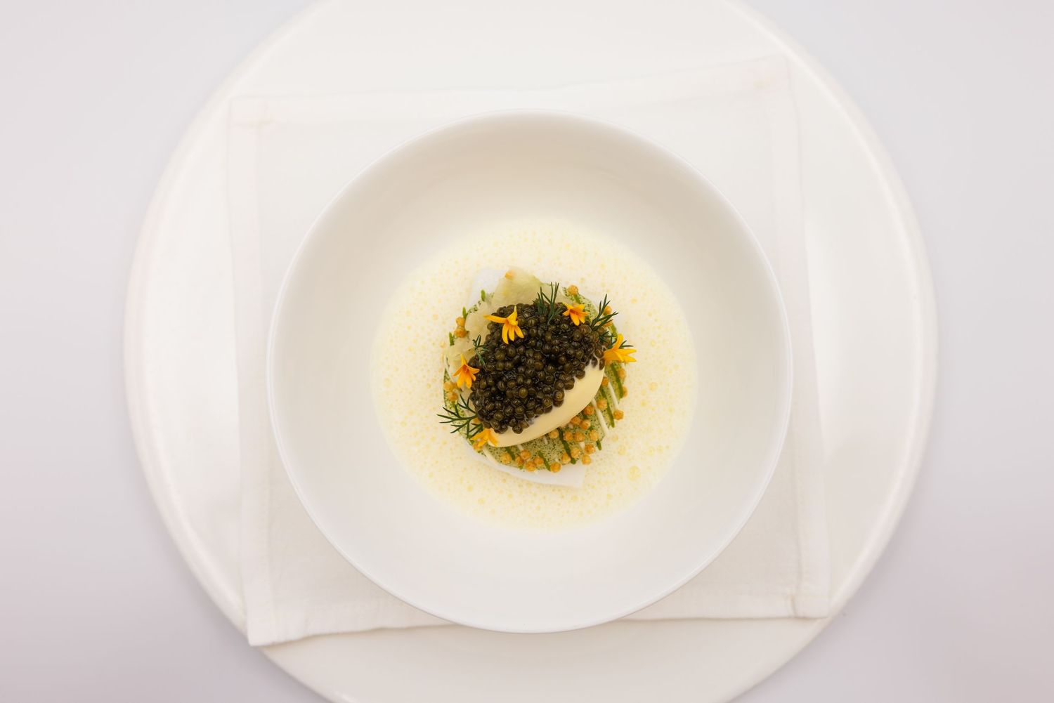 Elegant white bowl with caviar, herbs, and citrus garnish in a light broth, served on a white plate.