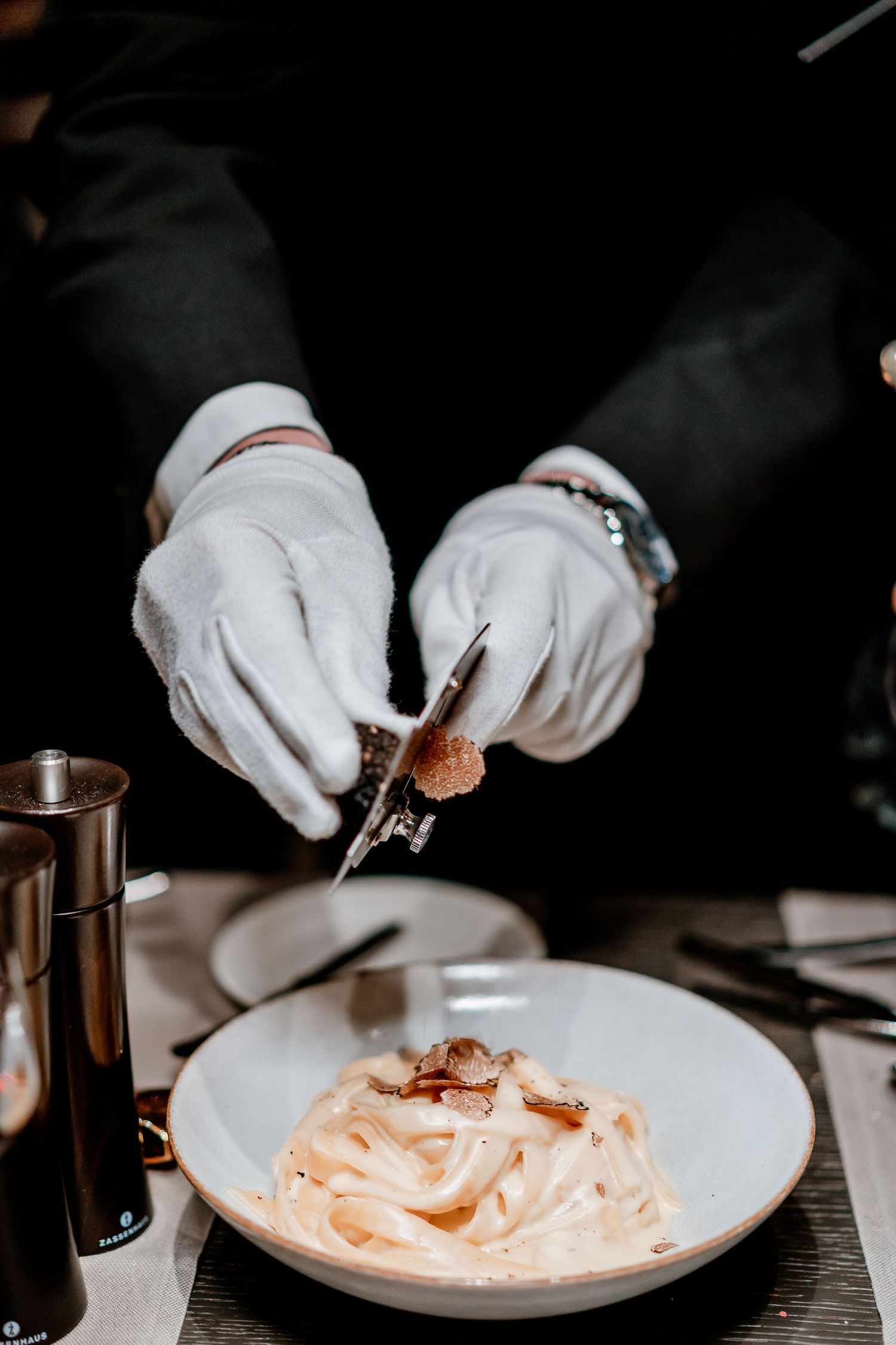 Gloved hands shaving fresh truffle over creamy fettuccine pasta in a white bowl at an upscale restaurant.
