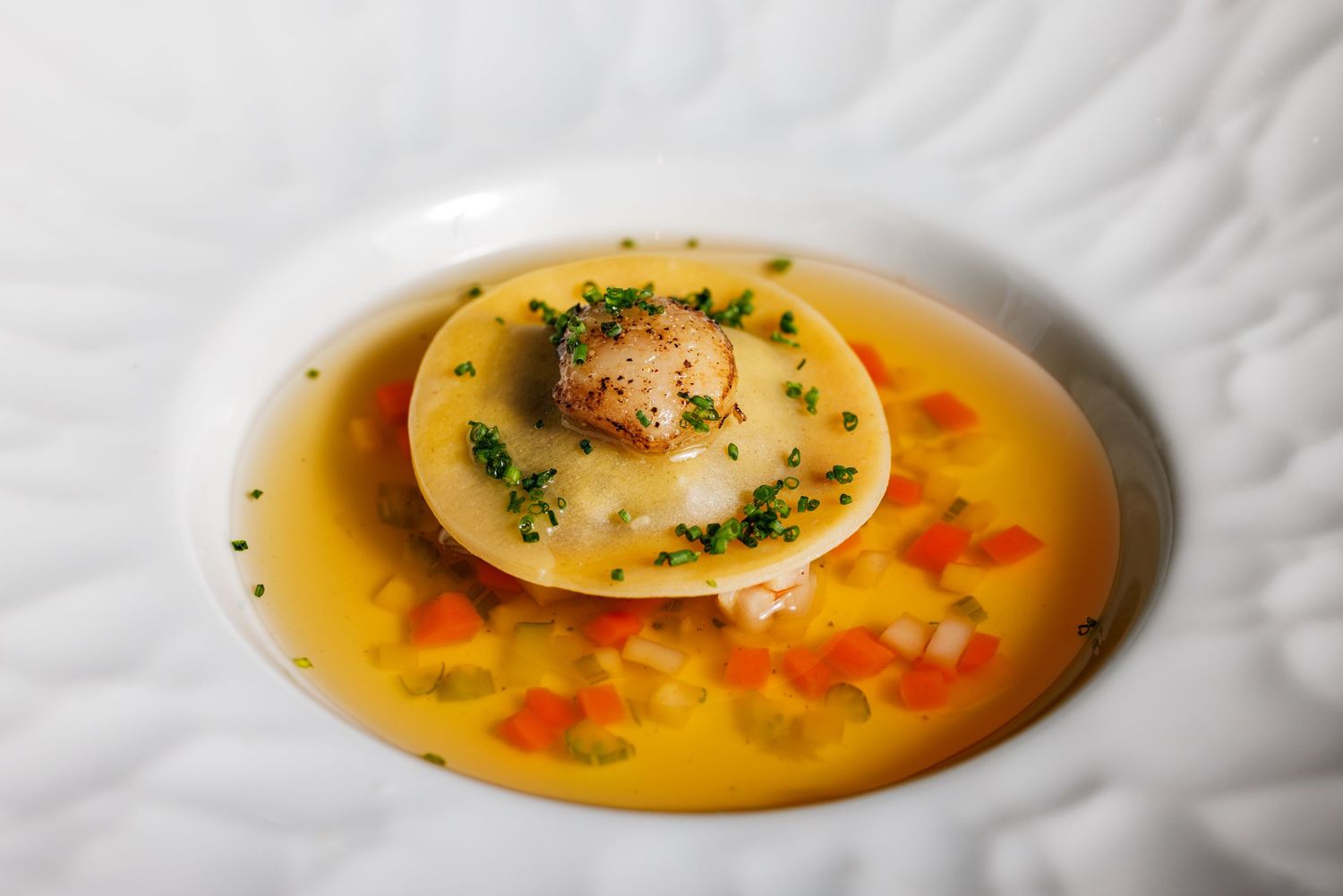 Seared scallop on pasta disc in golden broth with diced peppers and fresh herbs in white bowl.