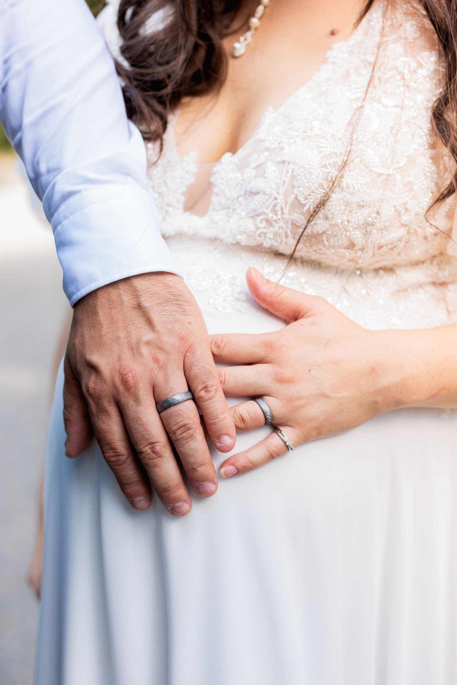 Newlywed couple holding hands showing silver wedding rings, bride in white lace dress, groom in light blue suit.