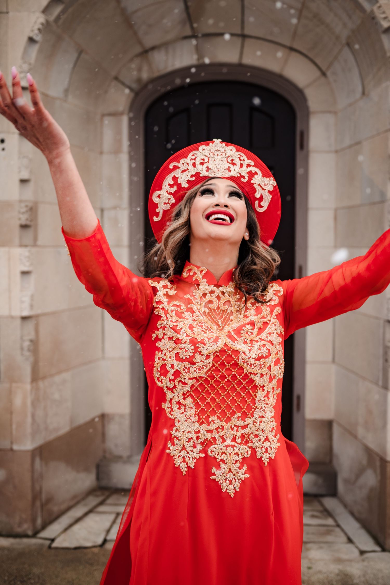 Person in vibrant red traditional dress with gold embroidery poses joyfully in stone archway corridor.