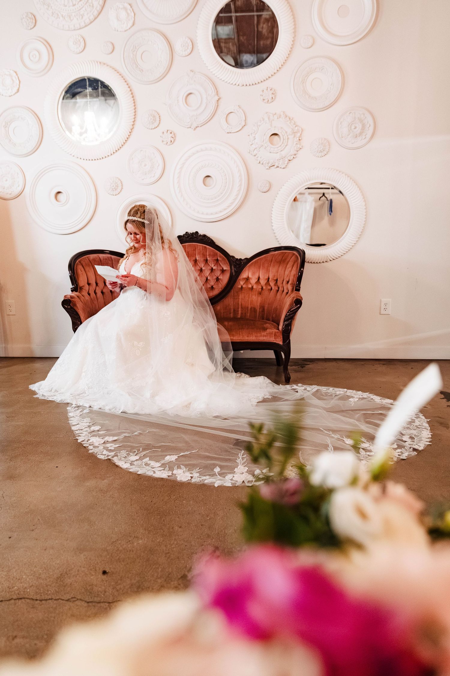 Elegant bridal portrait in white gown on vintage brown settee with decorative white plates adorning the wall behind.