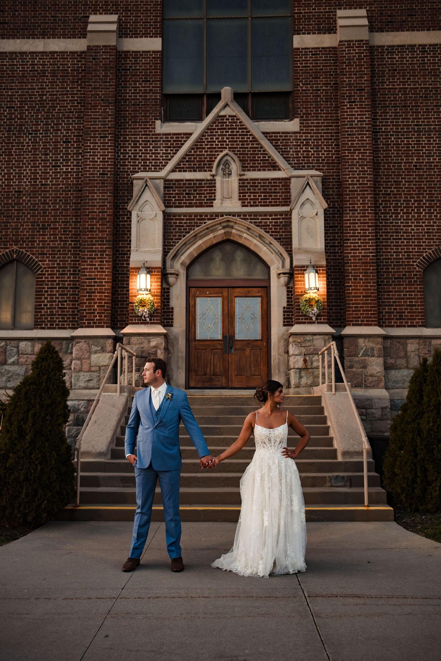 A couple in wedding attire stands holding hands in front of a grand brick church entrance with gothic architecture at dusk.