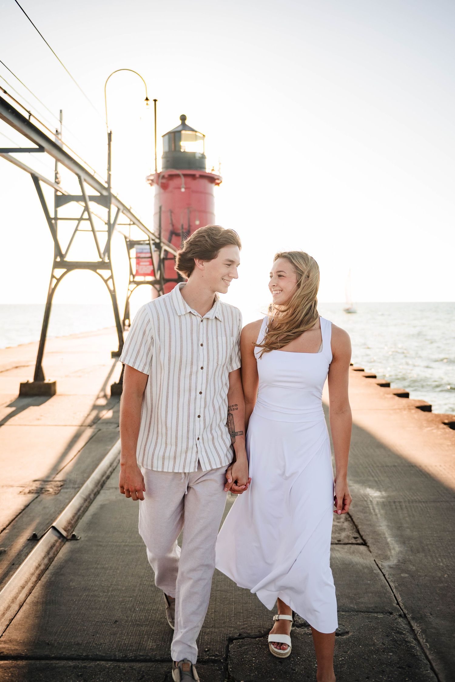 Couple walks hand in hand along pier with red lighthouse in background.